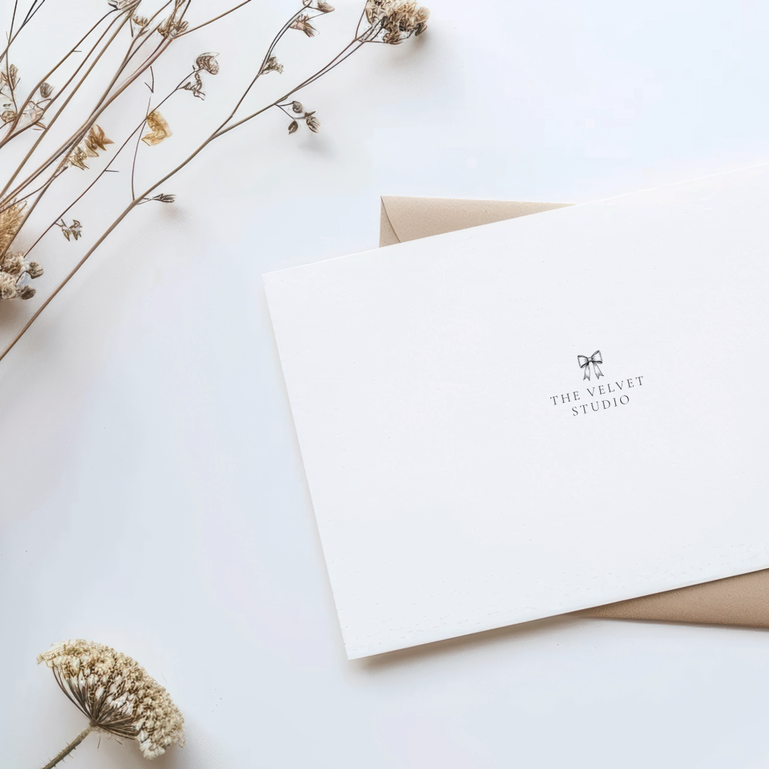 A white card with black text and a bow illustration, reading "The Velvet Studio," resting on top of a beige envelope on a white surface, with dried flowers nearby.