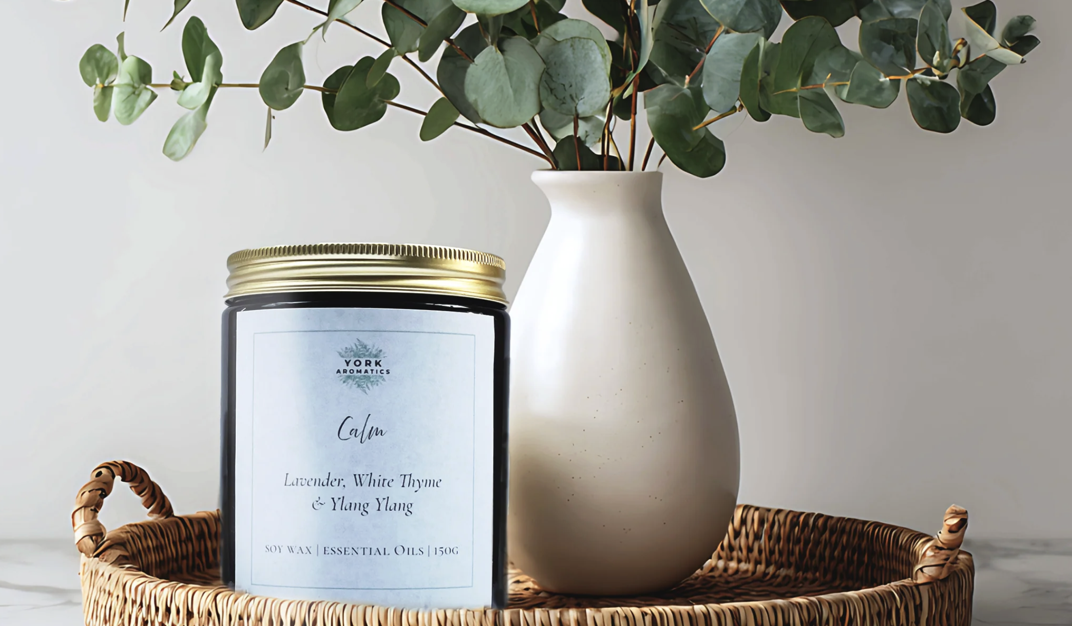 York Aromatics A black candle jar labeled 'Calm' with lavender, white thyme, and ylang ylang, placed on a woven basket tray alongside a white ceramic vase with green eucalyptus leaves, all set on a white surface against a plain background.