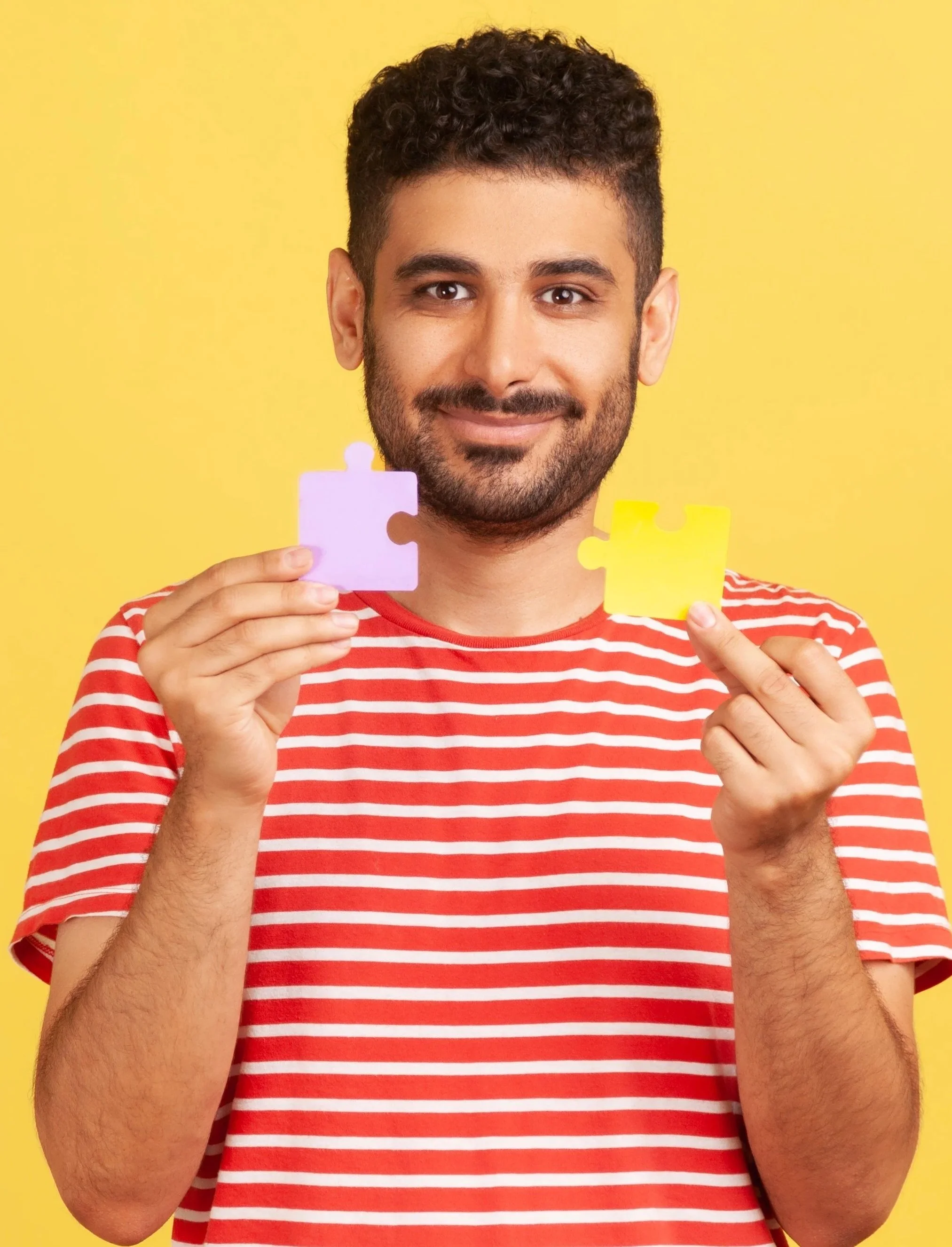 Man met donker haar en baard houdt een paar puzzelstukjes vast, een roze en een geel, tegen een gele achtergrond.