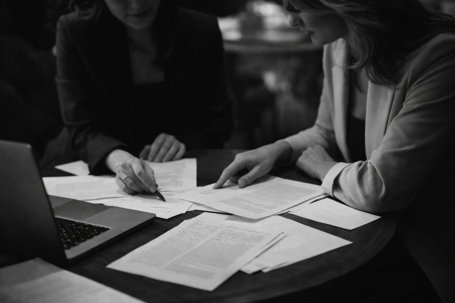 Two women are reviewing and discussing multiple documents and papers spread out on a table, with a laptop nearby, in a black-and-white photo.