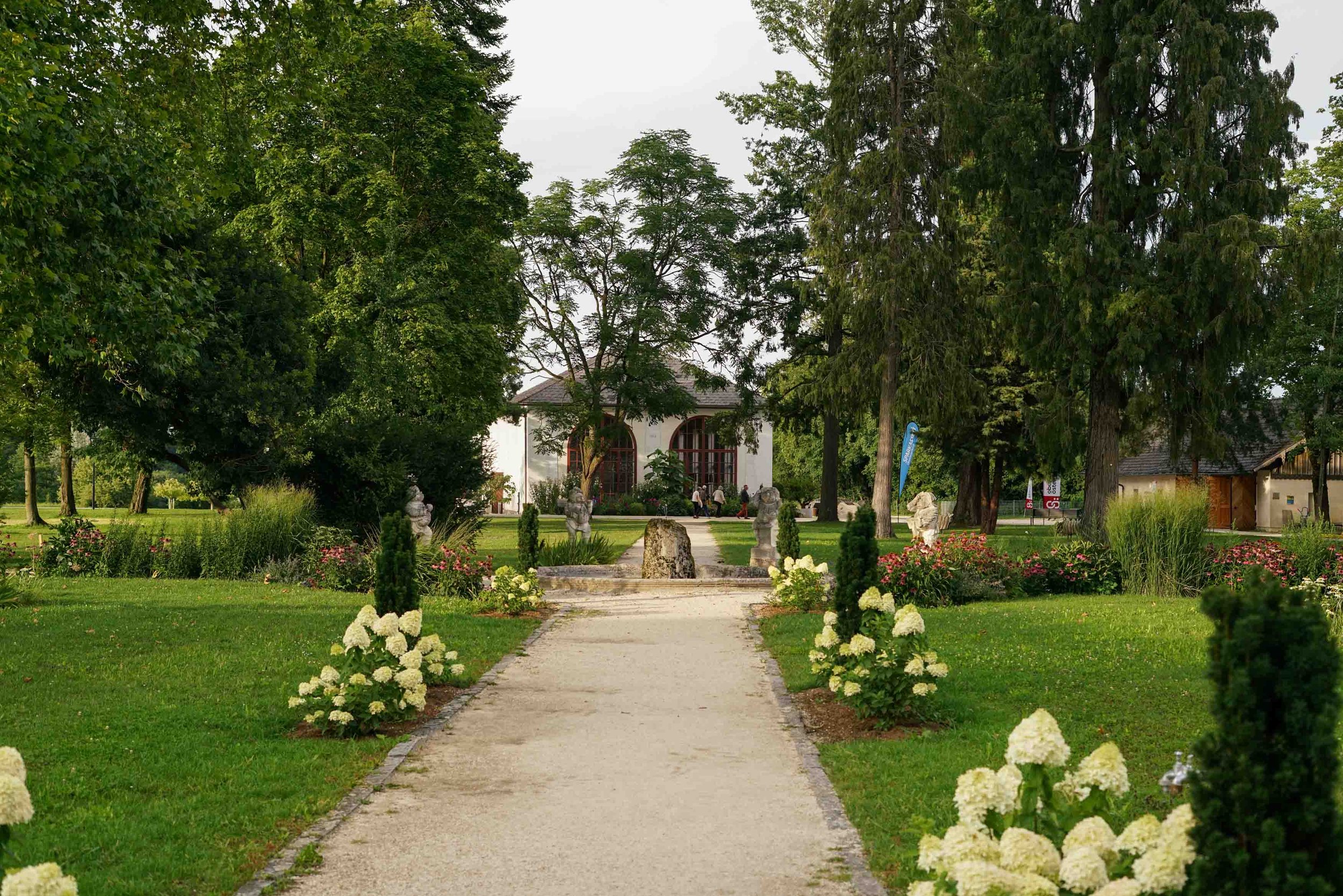 Ein gepflegter Garten mit einem gepflasterten Weg, umgeben von Blumen und Bäumen, mit einem weißen Gebäude im Hintergrund.