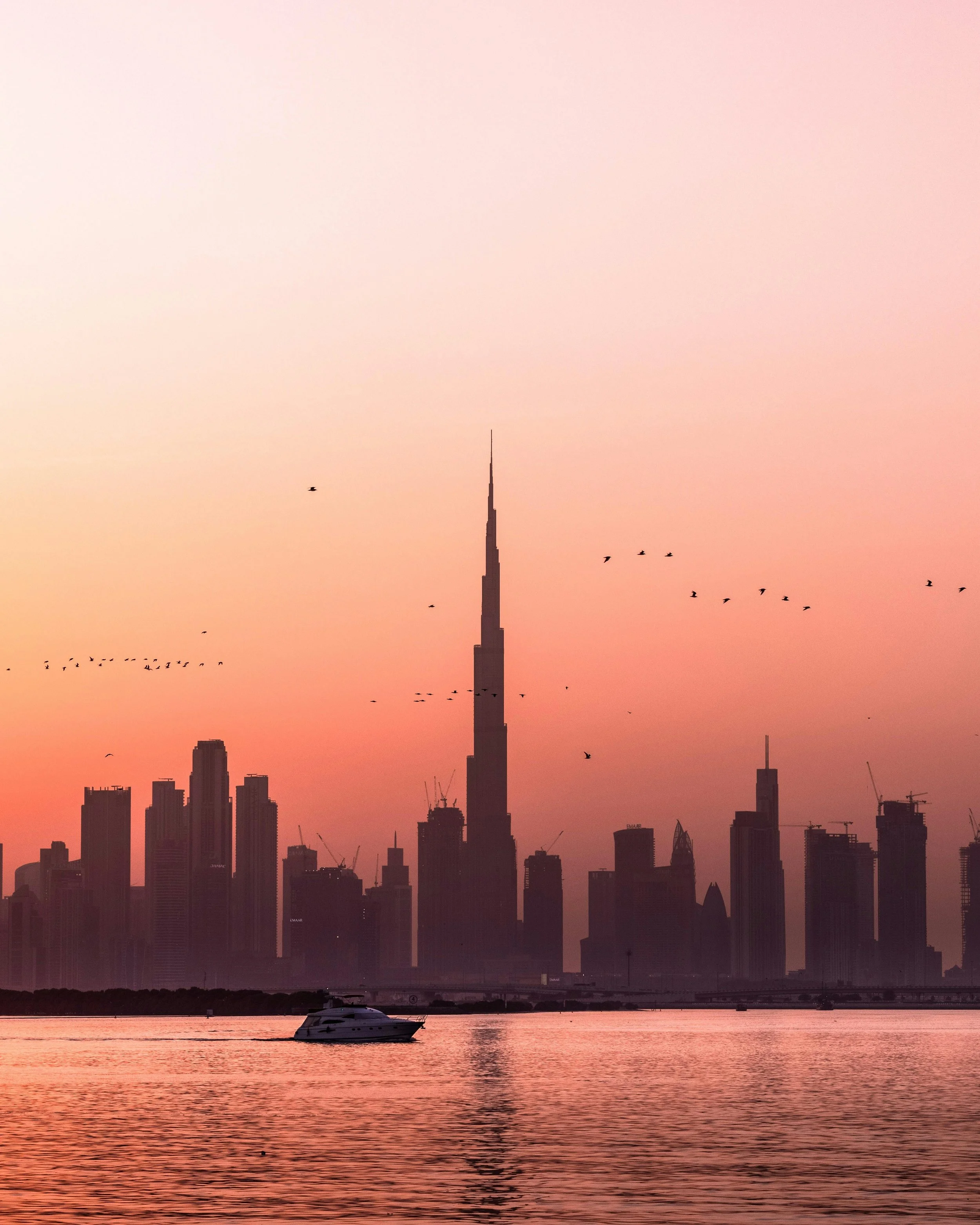 Sunset over Dubai skyline with the Burj Khalifa, a yacht on the water, and birds flying in the sky.