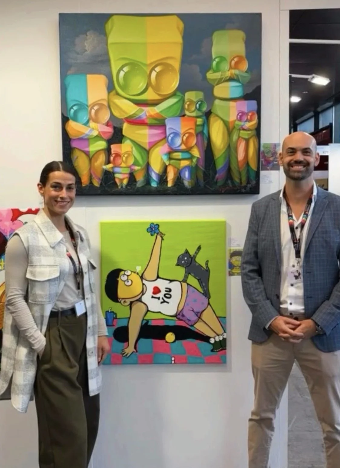 Two people standing in front of colorful artwork at an art exhibit, with a woman on the left and a man on the right, both smiling.