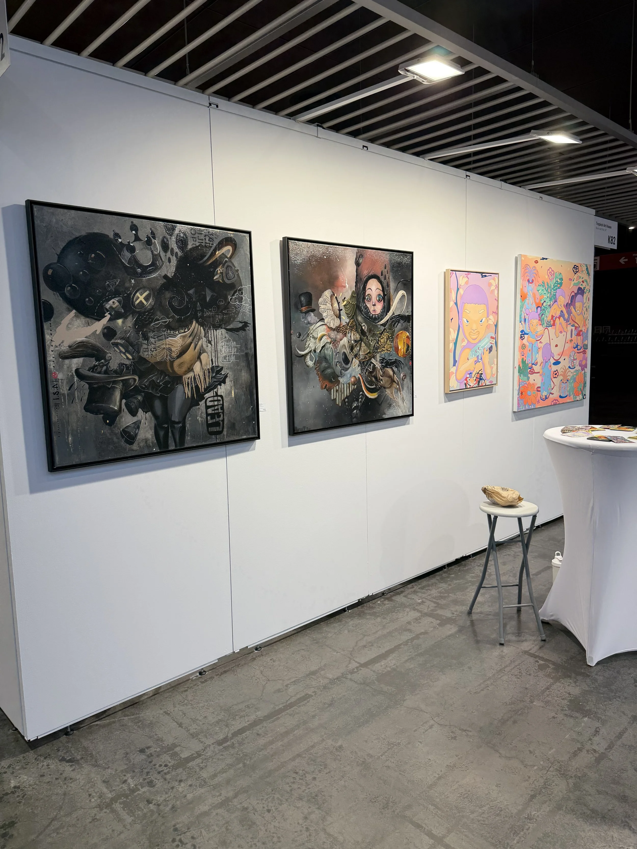 Four colorful paintings displayed on a white gallery wall. The first two are dark, abstract pieces with figures and intricate designs. The third shows a stylized portrait of a woman with purple hair. The fourth depicts a vibrant, fantasy scene with v