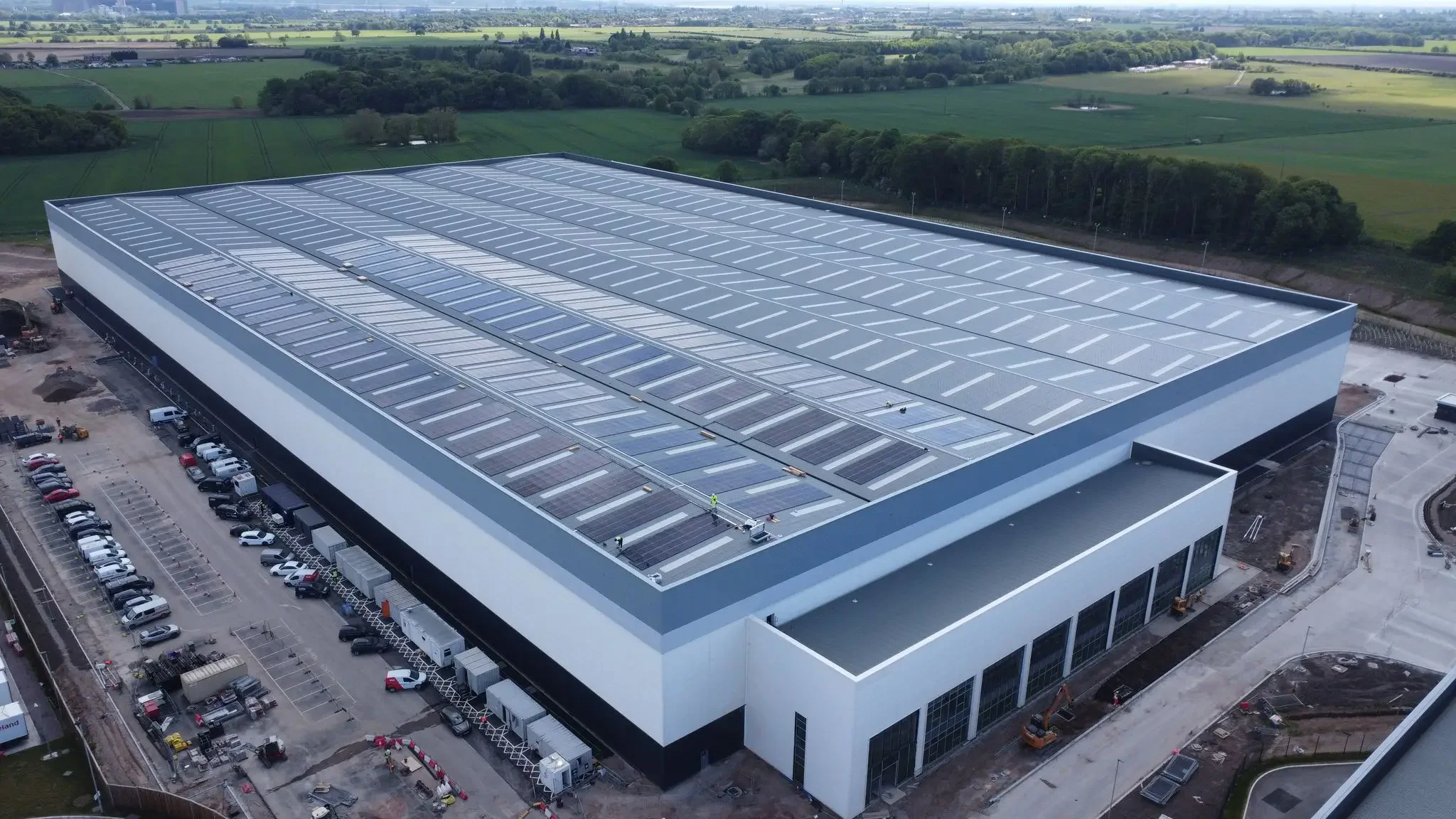 An aerial view of a large building with solar panels on the roof, surrounded by parking lots and construction areas, with green fields and trees in the background.