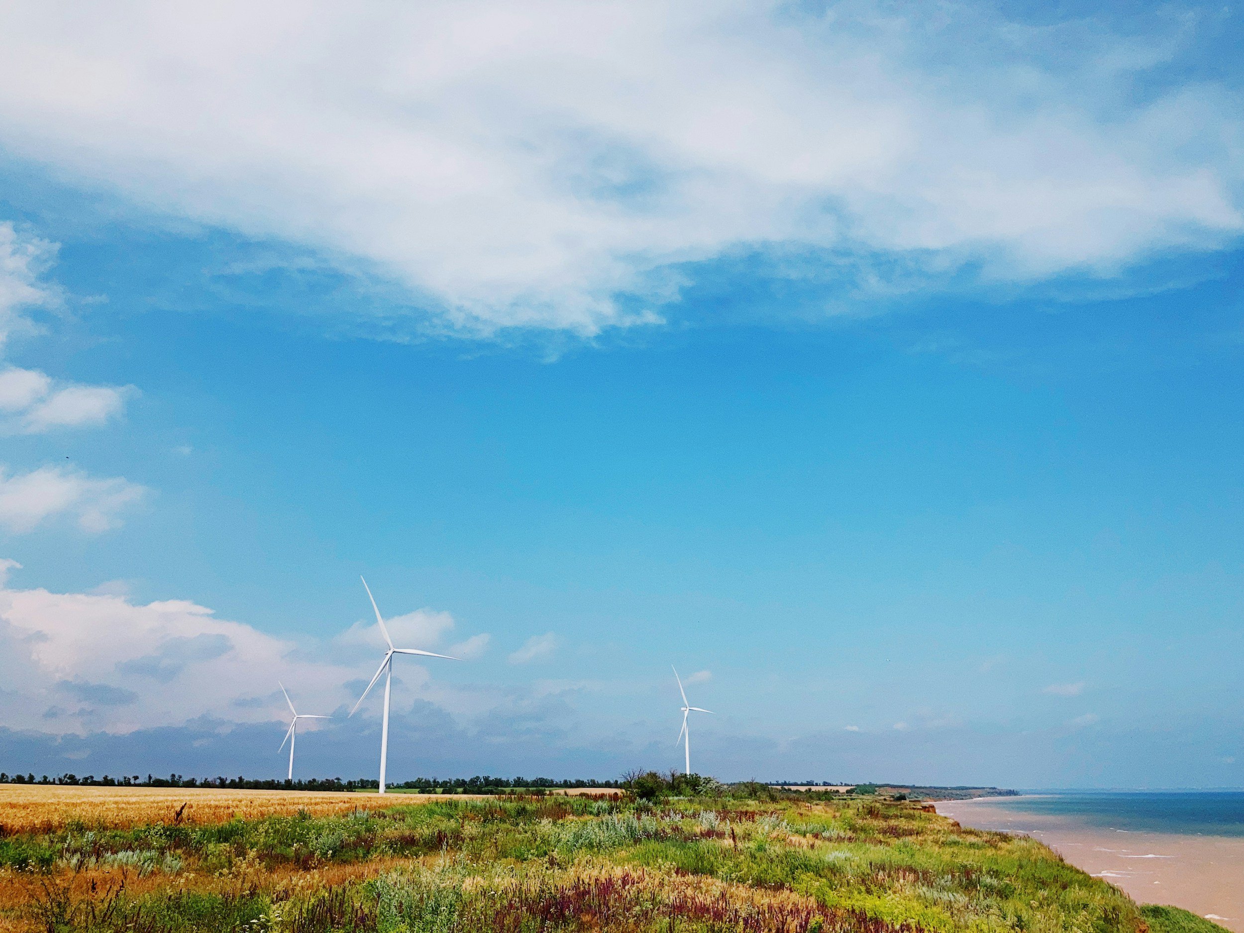 A coastal landscape with grassy dunes, three wind turbines, a sandy beach, and a partly cloudy blue sky.