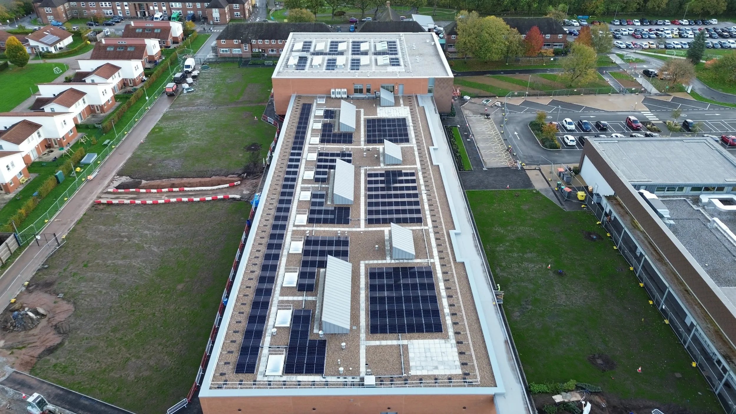 Aerial view of a flat commercial building roof with multiple solar panel installations, surrounded by grass, parking lots, and residential houses.