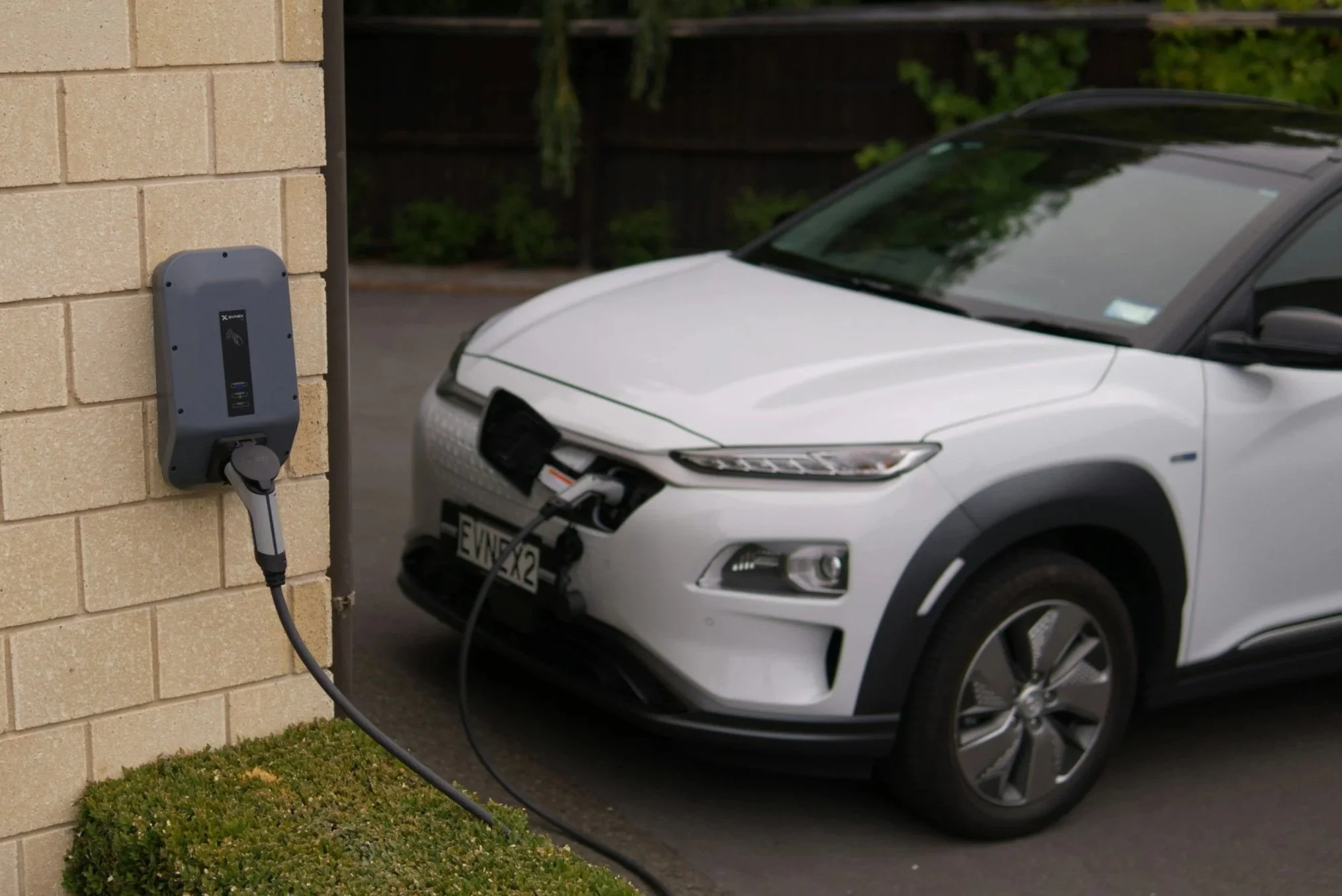A white electric vehicle is plugged into a charging station on a brick wall, with a garden background.