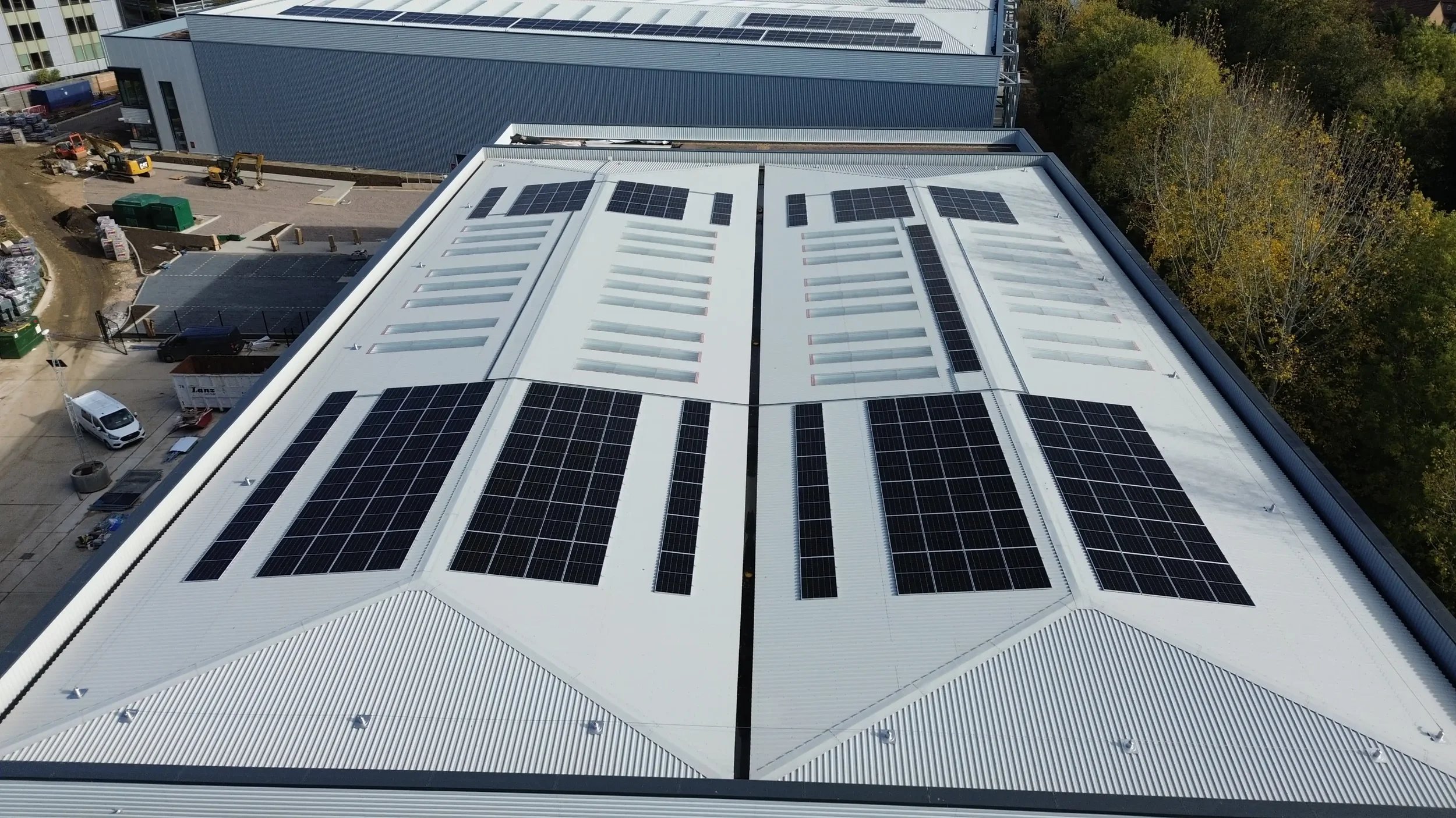 Aerial view of a large white commercial building rooftop with multiple solar panels installed on different sections, surrounded by trees and a parking area with construction equipment.