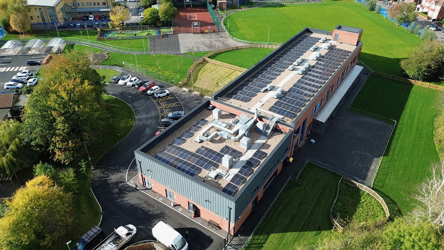 An aerial view of a building with a large roof covered in solar panels, surrounded by parking lots, green fields, trees, and sports courts.