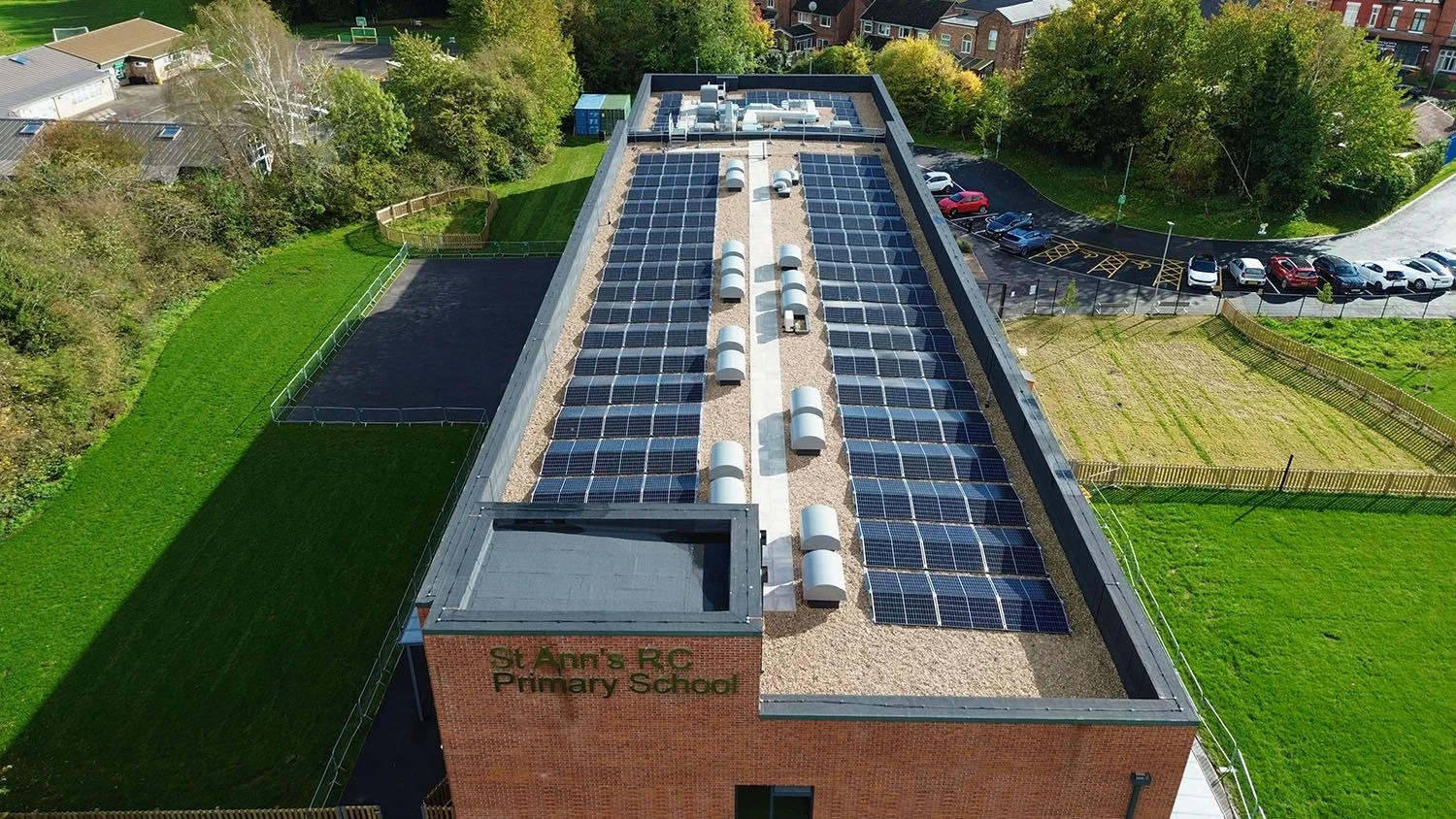 Aerial view of St Ann's RC Primary School building with solar panel installation on the roof and surrounding green fields and trees.