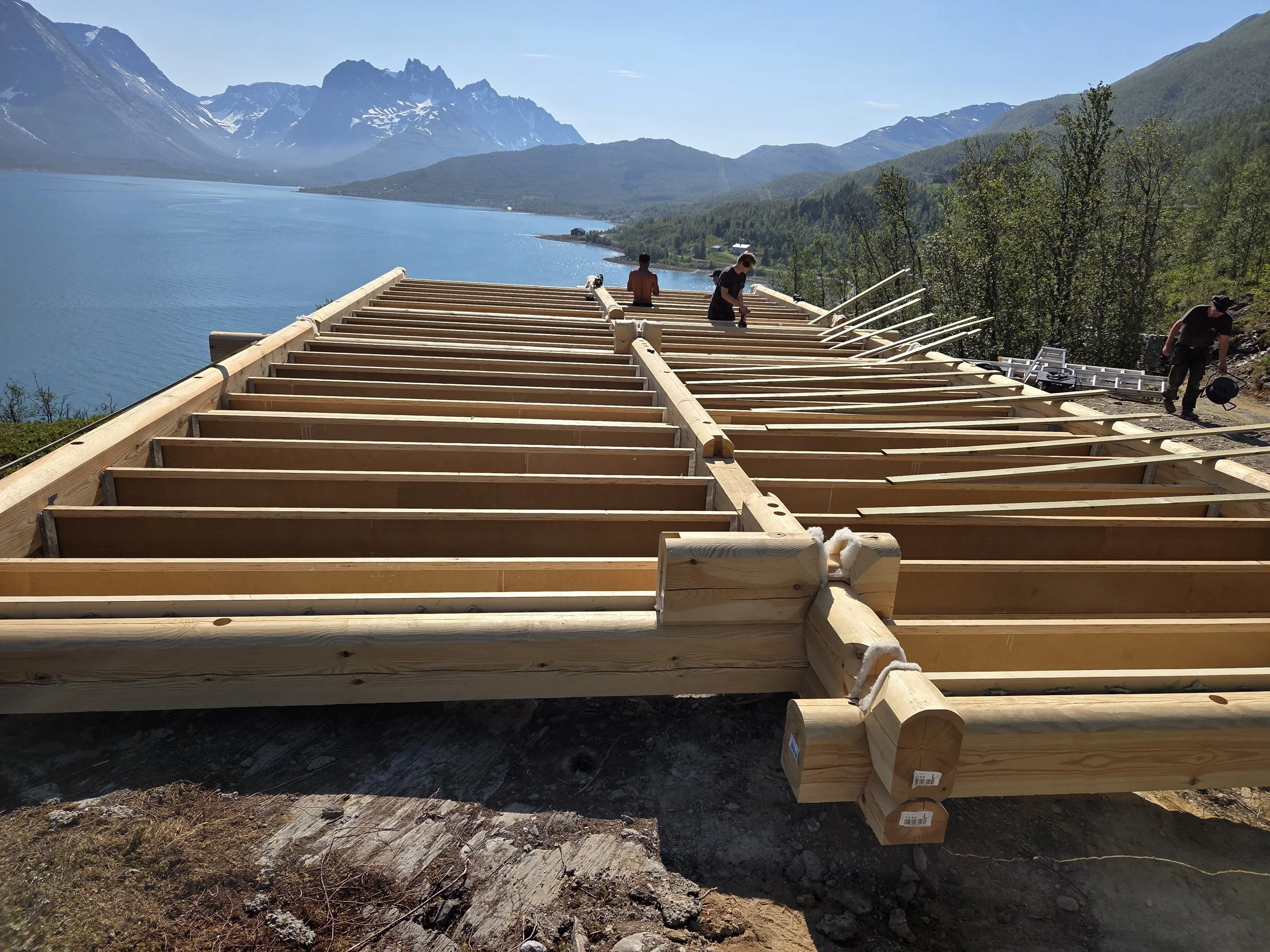 Construction workers building a wooden dock or deck next to a large body of water, with mountains and trees in the background.