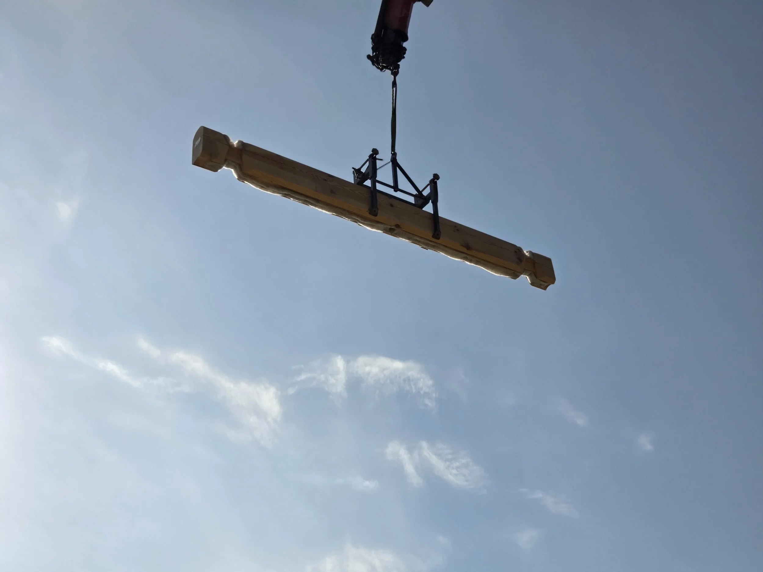 A crane lowering a large wooden beam against a blue sky with some clouds.