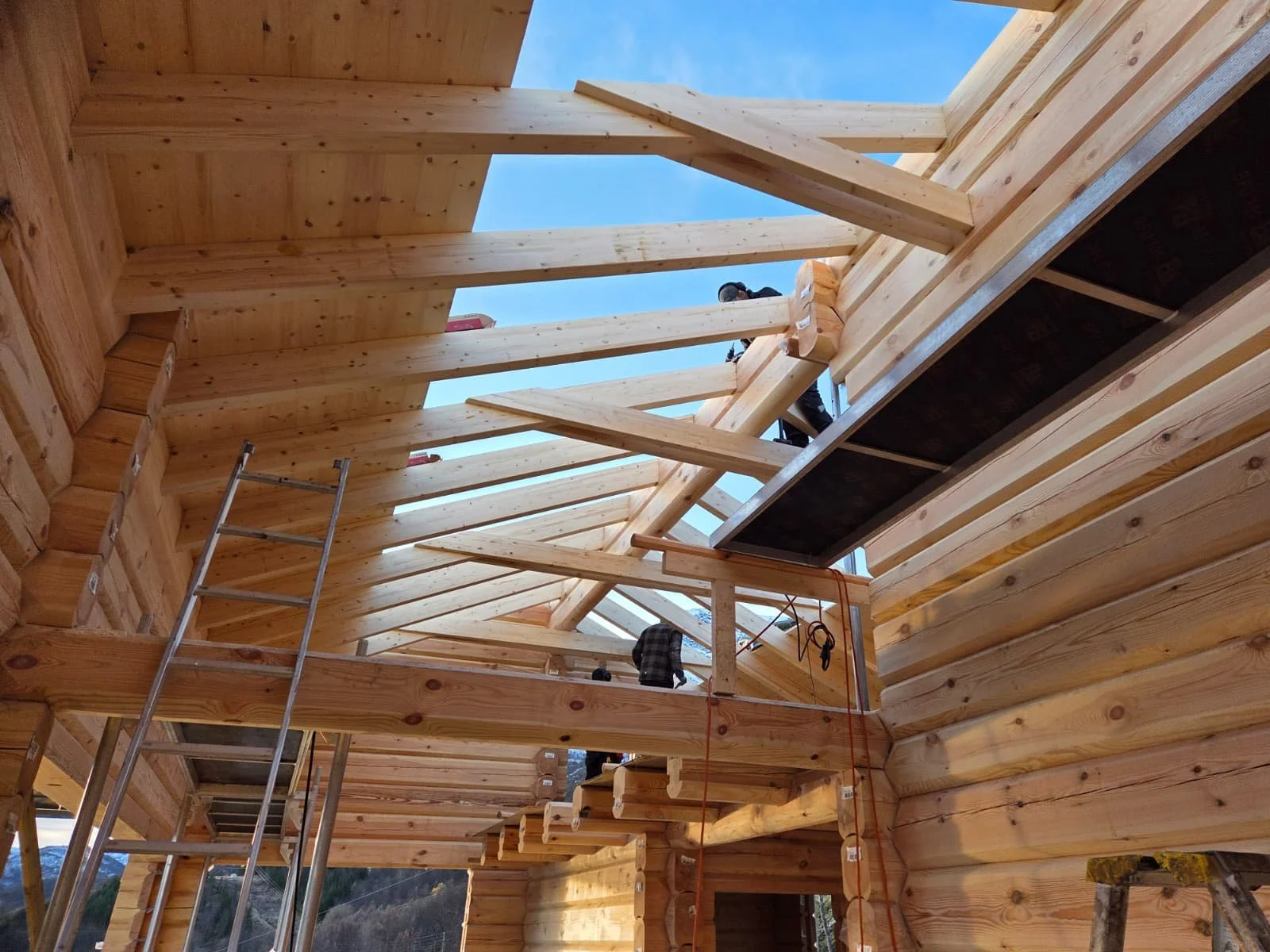 Wooden house under construction with an open roof and workers installing roof beams, ladders leaning against the walls, and clear blue sky visible above.