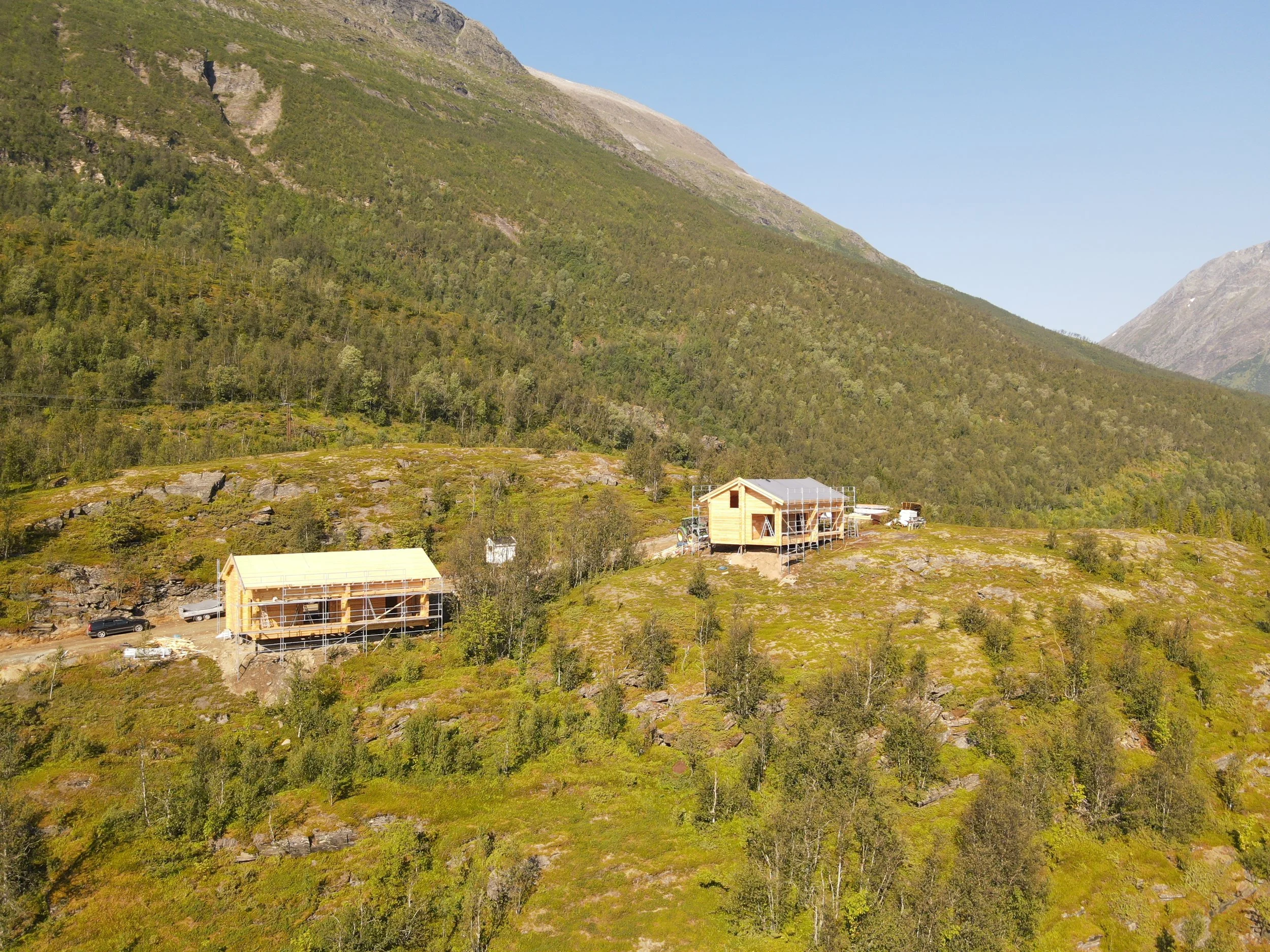 Two wooden houses under construction on a hillside surrounded by green trees and mountains.