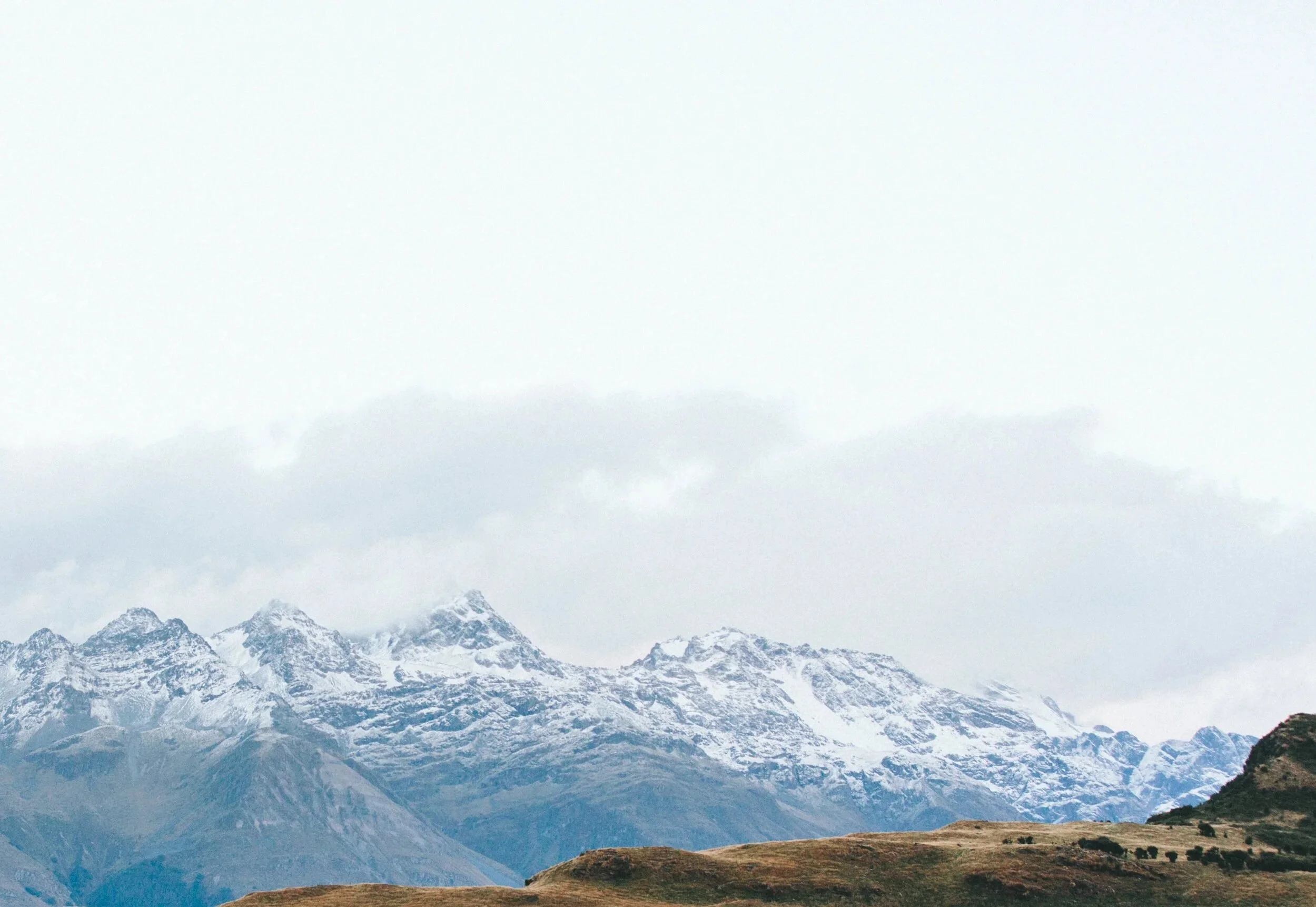 Snow-capped mountains in the background with rolling brown hills in the foreground under a cloudy sky.