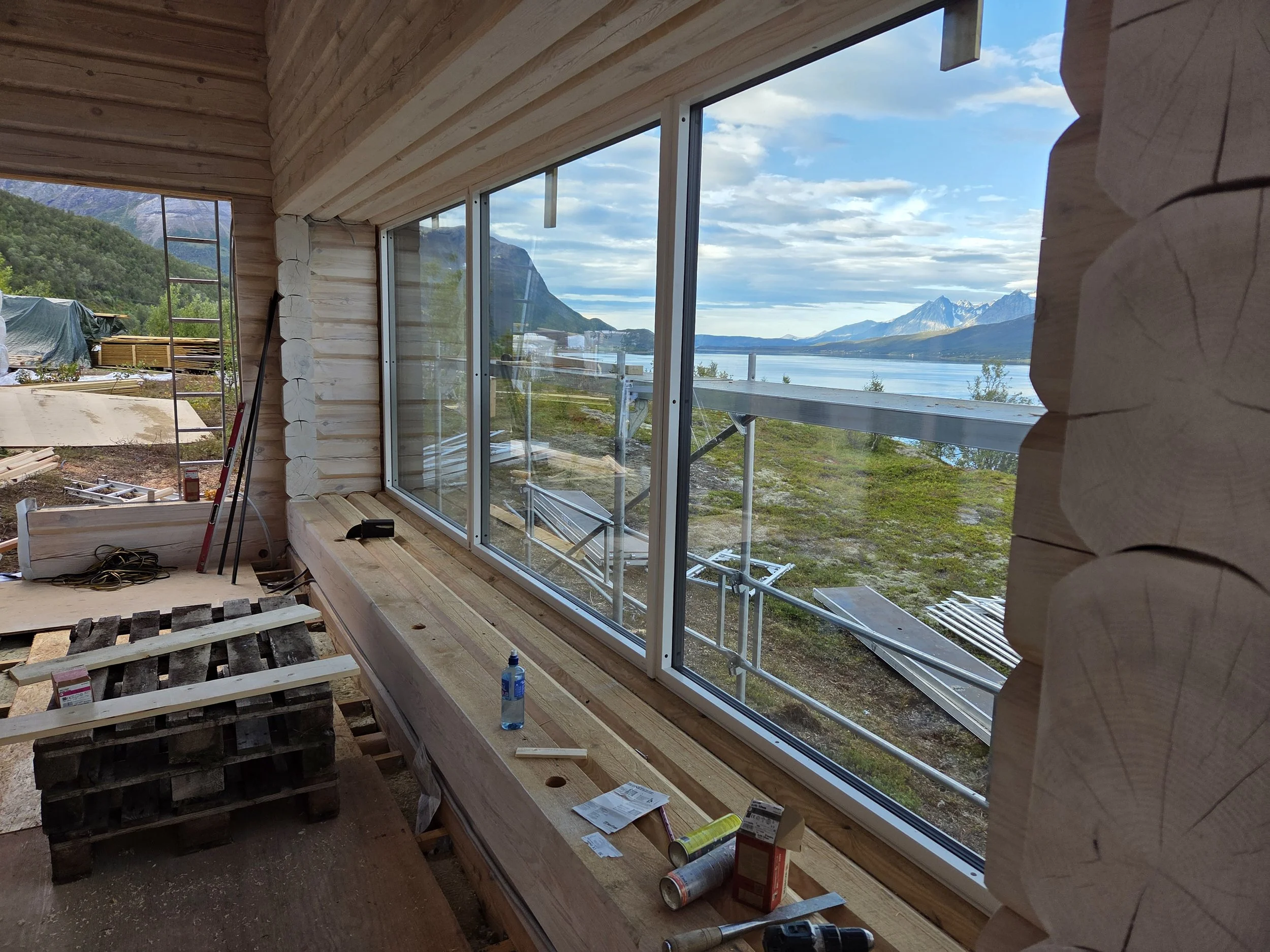 Interior view of a log cabin under construction, with a large window overlooking a scenic landscape of mountains, water, and sky.