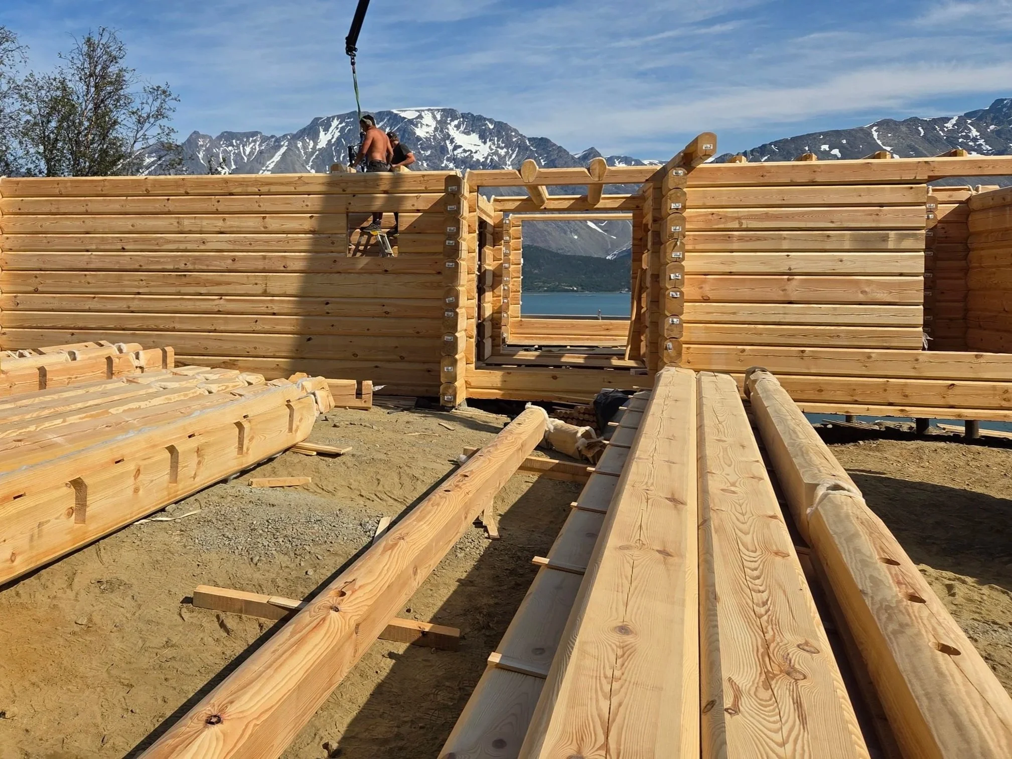 Under construction wooden house with mountains in the background, showing workers framing the walls, with large wooden beams and planks on the ground.