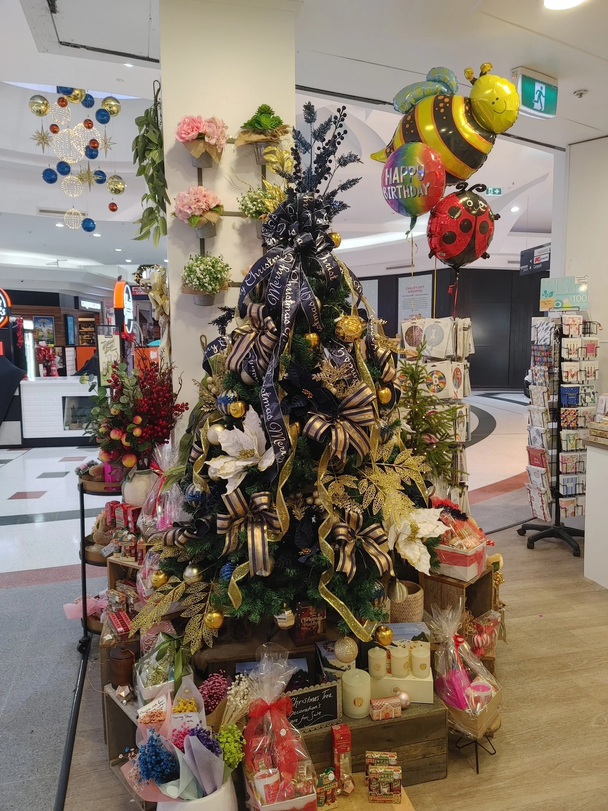Decorated Christmas tree with gold, black, and white ribbons, gold ornaments, and poinsettias, surrounded by gift baskets and wrapped presents, with balloons including a bee, ladybug, and “Happy Birthday” message, in a shopping mall.