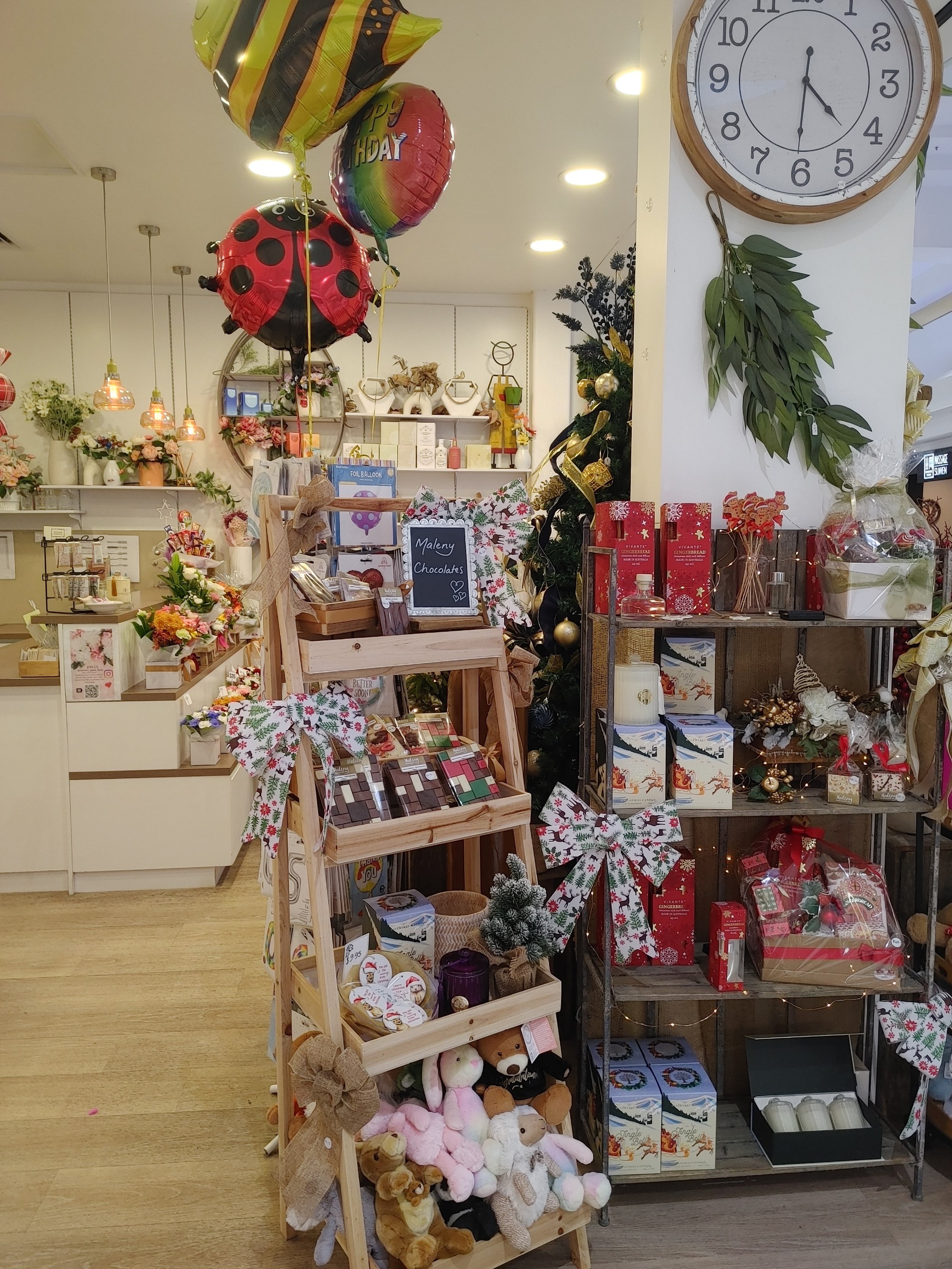 Interior of a store decorated for Christmas with balloons, plush teddy bears, holiday gifts, and a large round clock on the wall showing 6:30.