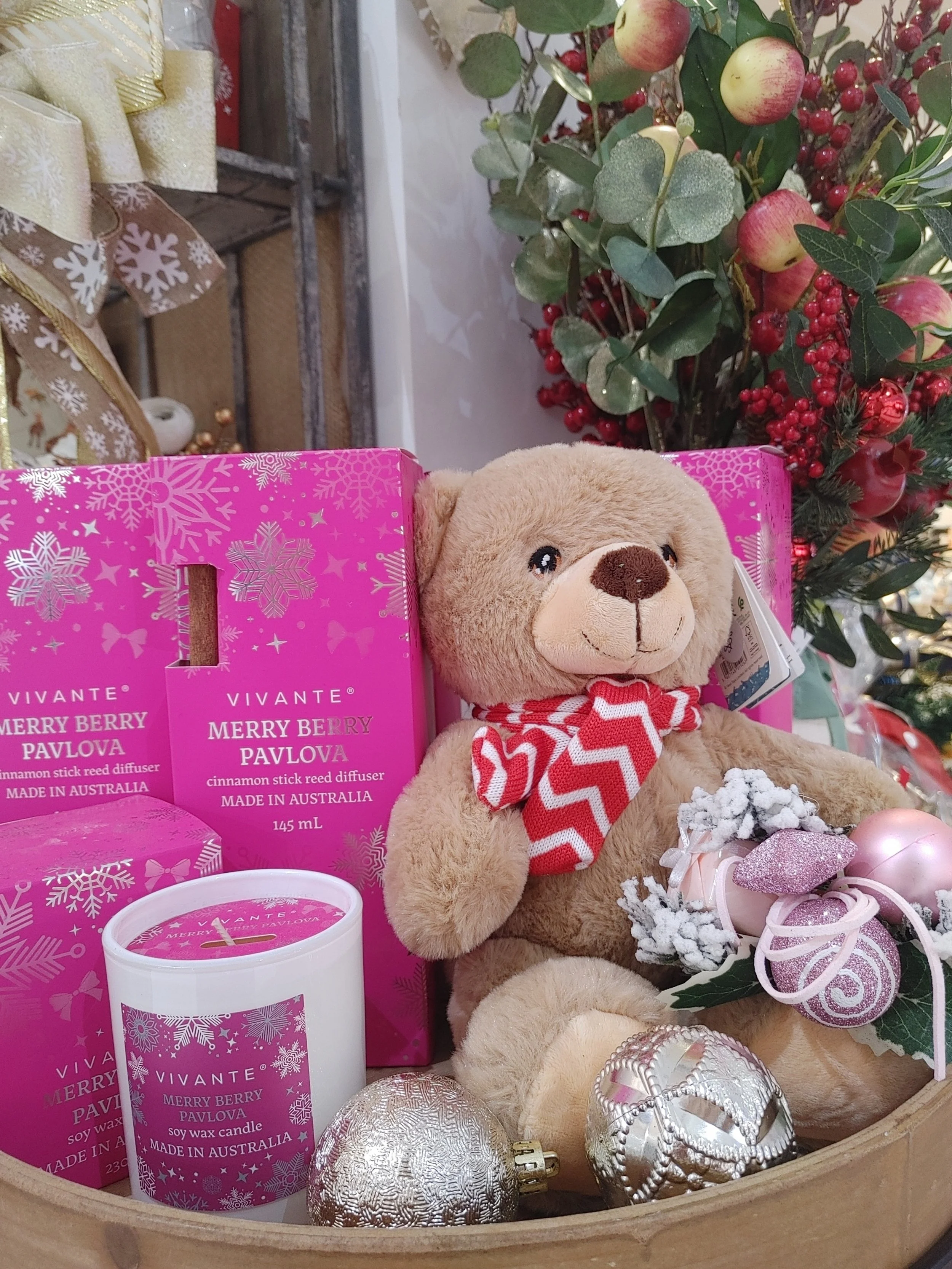 A Christmas display featuring a plush teddy bear with a red and white chevron scarf, surrounded by pink and silver ornaments, a candle, and Merry Berry Pavlova cinnamon stick reed diffuser boxes and a pink candle, with Christmas decorations in the ba