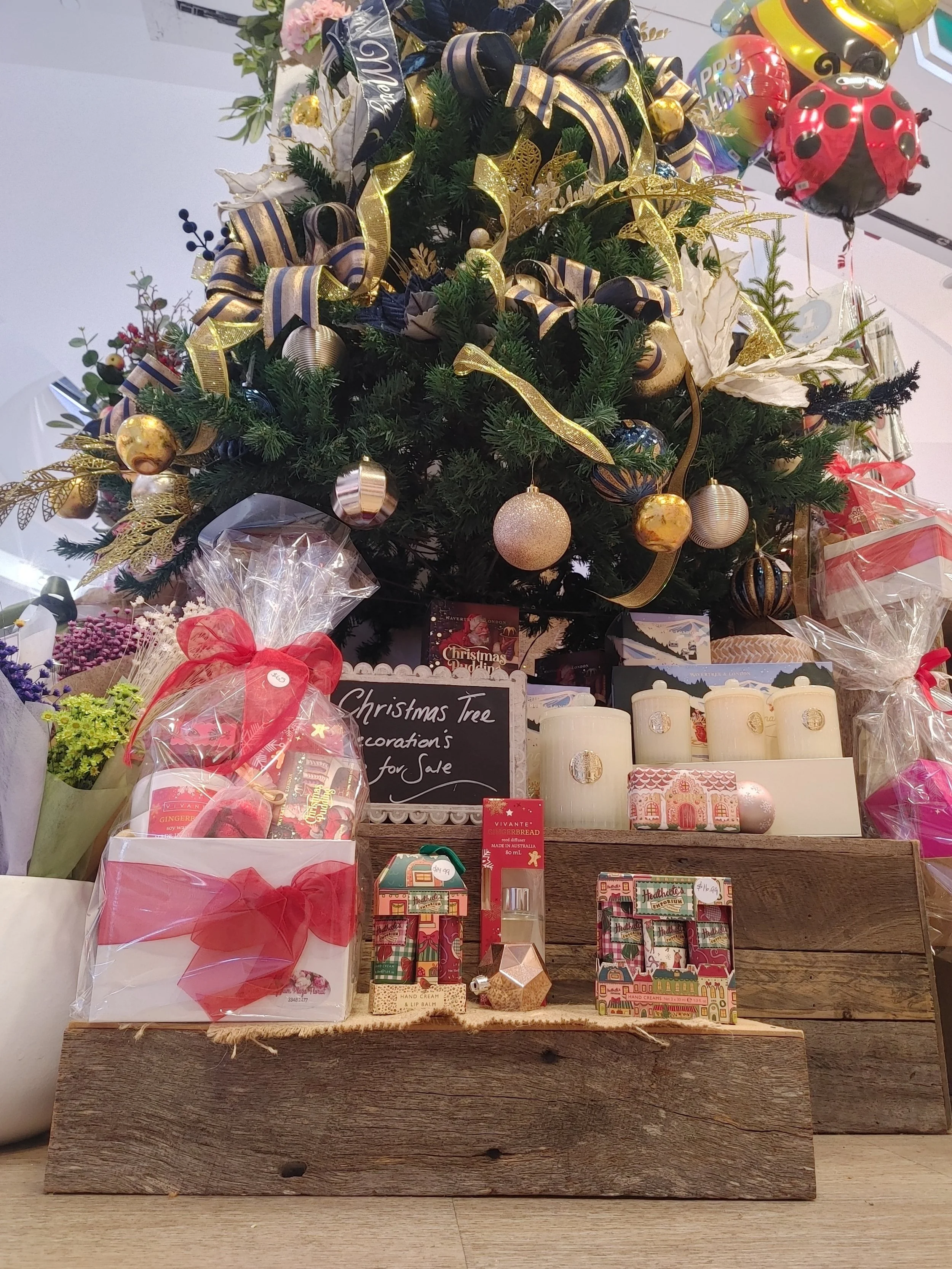 Decorated Christmas tree with gold, black, and white ribbons, gold ornaments, and gold ribbon bows. Presents and gifts are placed around the base, some wrapped with red ribbons, and there are candles, small boxes, and a chalkboard sign reading 'Chris
