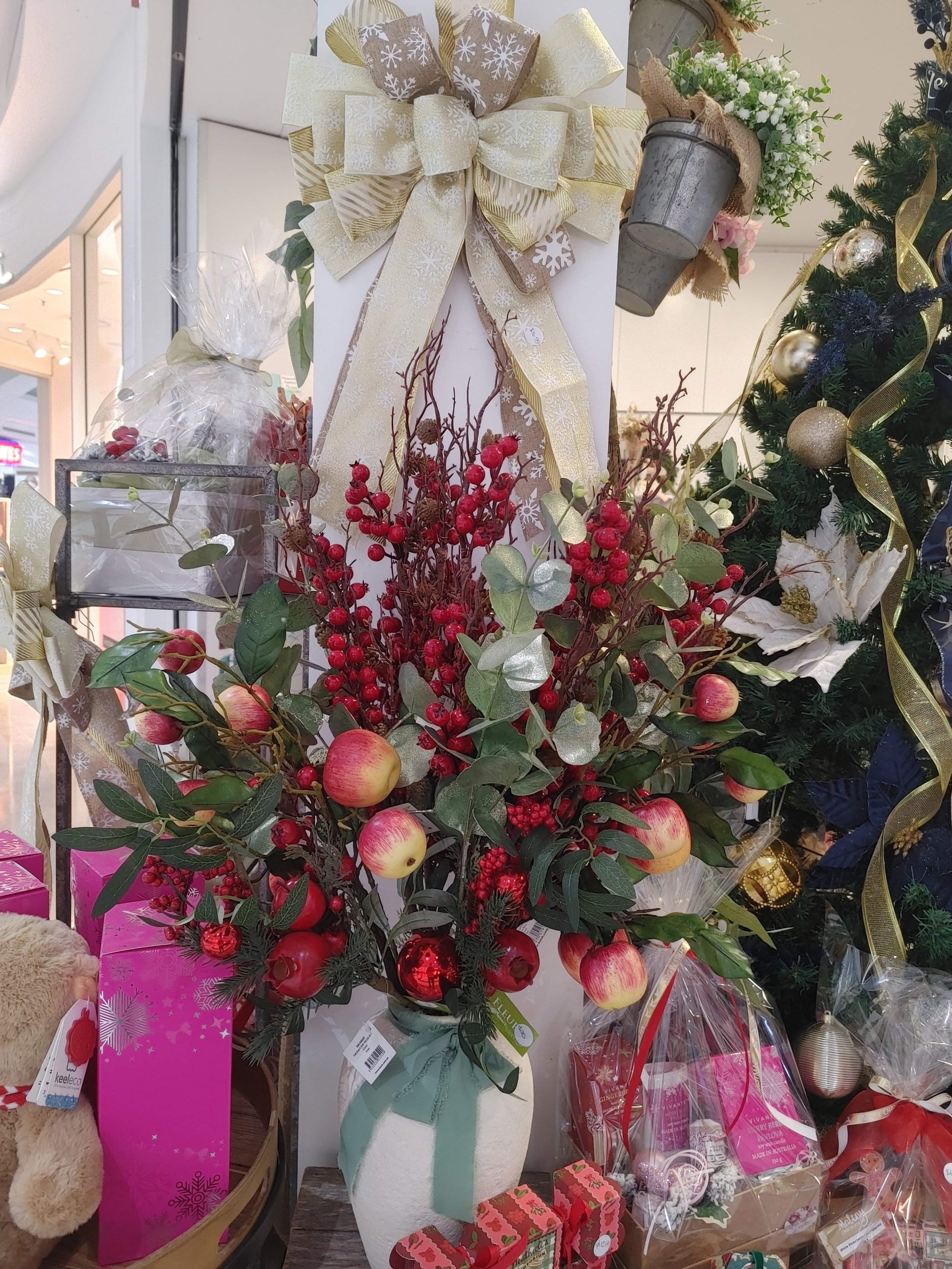 A Christmas floral arrangement with red berries, apples, and green leaves in a white vase, adorned with a green ribbon, displayed among holiday decorations and gifts.