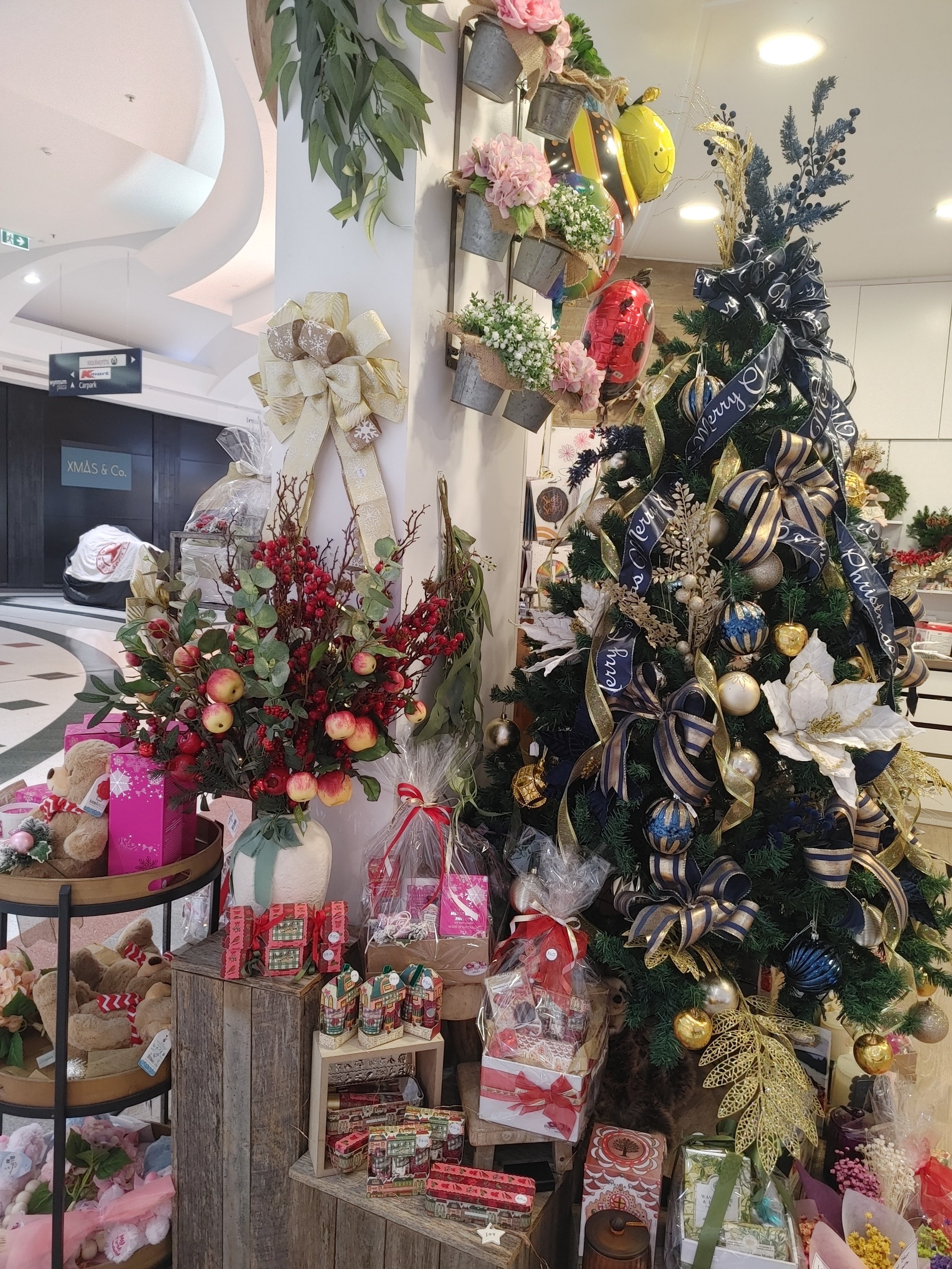 A decorated Christmas tree with gold, blue, and white ornaments, ribbons, and a 'Merry Christmas' bow inside a store. Surrounding the tree are gift boxes, wrapped presents, and holiday floral arrangements including red berries, apples, and pink and w