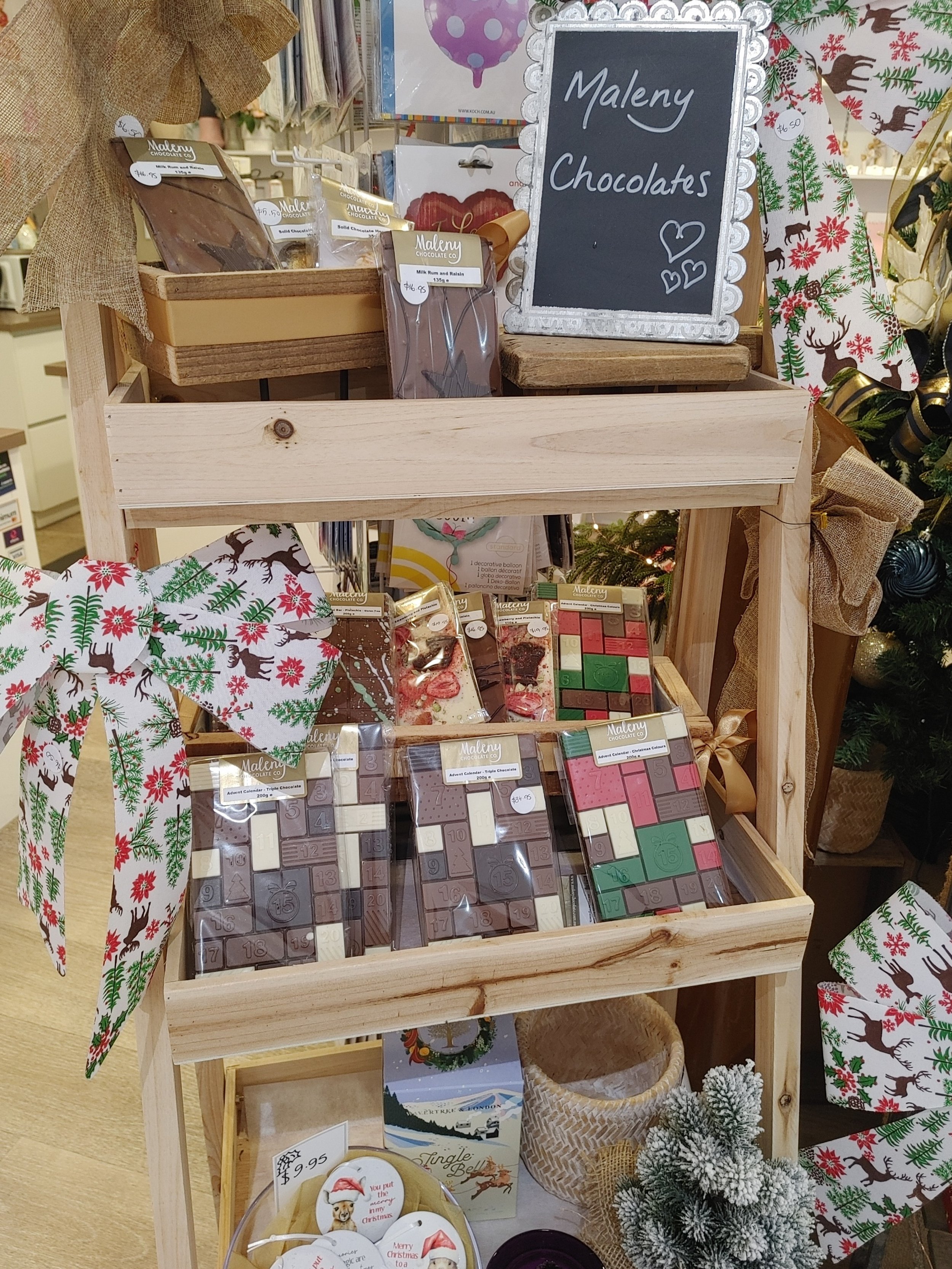 Display of Maleny chocolates and holiday-themed items on a wooden stand, with a chalkboard sign that reads 'Maleny Chocolates', decorated with Christmas ribbons, bows, and ornaments.
