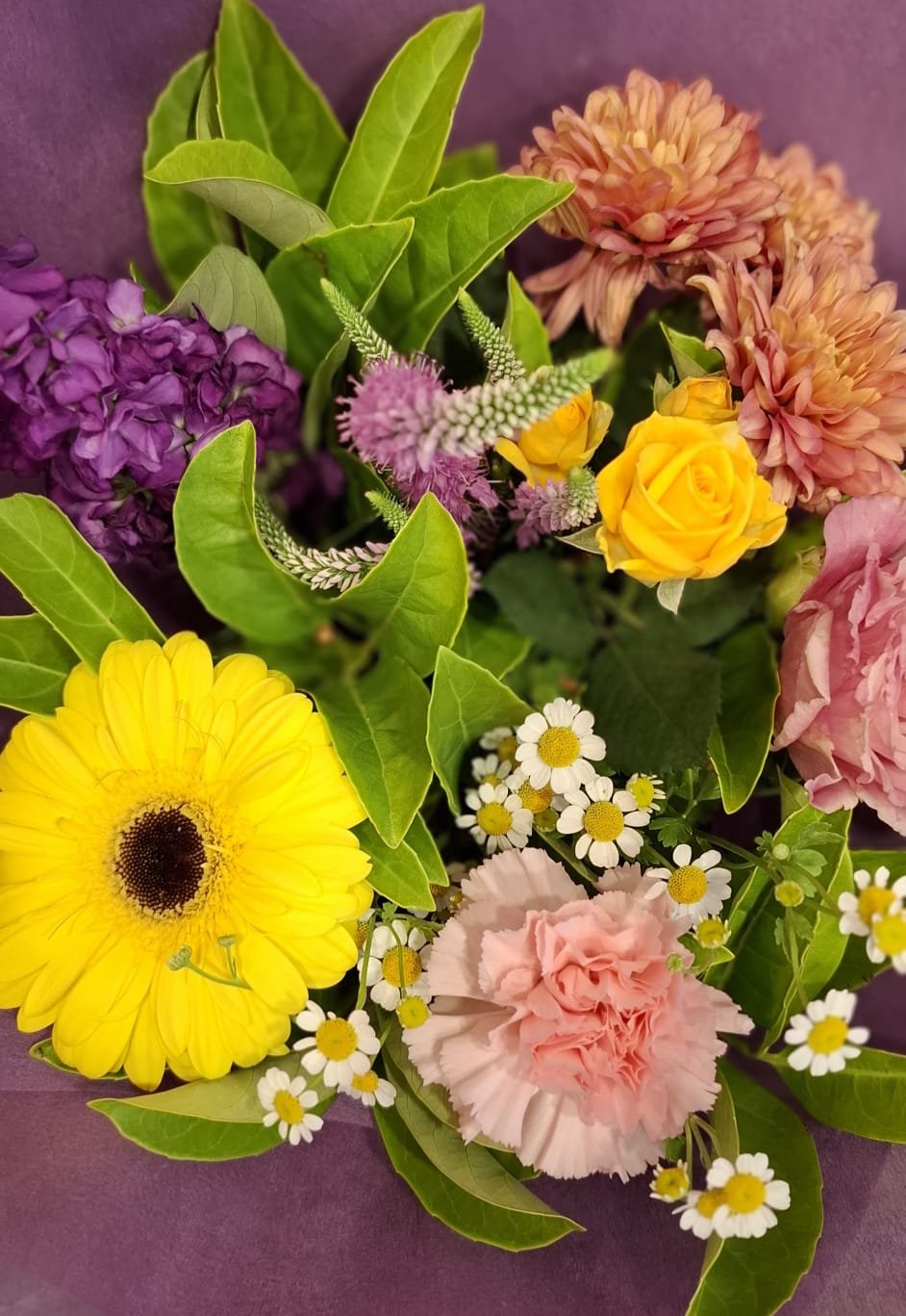 A colorful bouquet of flowers including yellow gerbera daisy, pink carnations, purple hydrangea, orange dahlias, small white daisies, and green leaves.