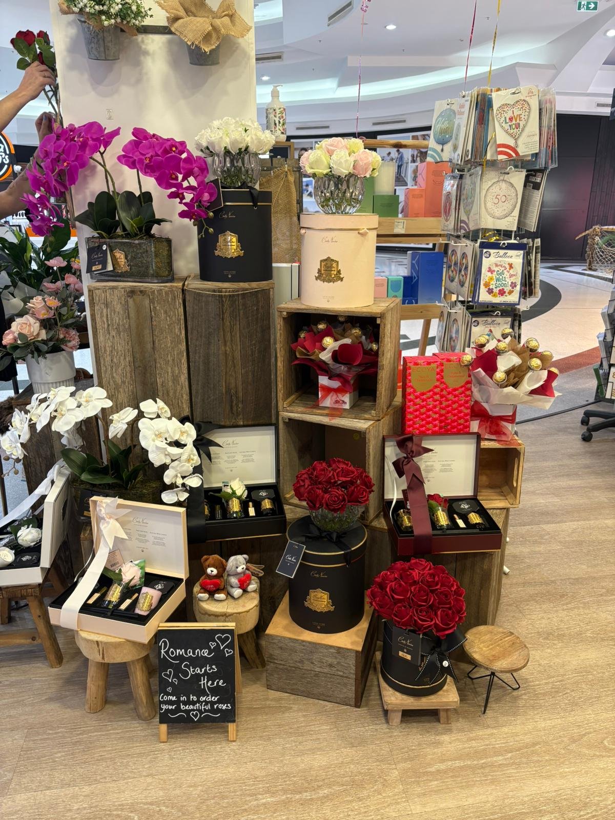 Display of various flower arrangements with roses and orchids, gift boxes, teddy bears, and greeting cards at a flower shop or mall.