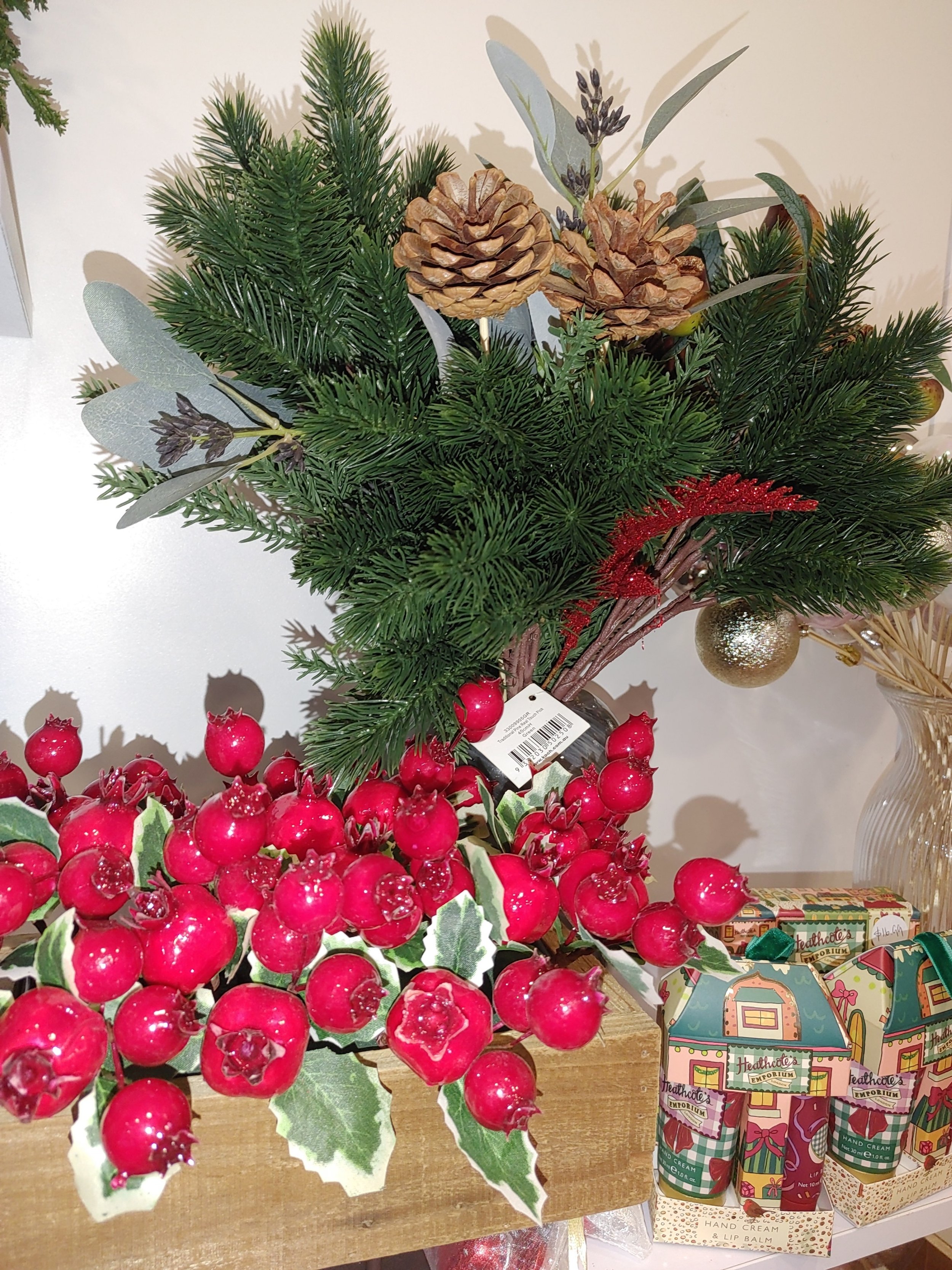 Christmas arrangement with pine, pinecones, red berries, and decorative green foliage, next to holiday-themed hand cream and lip balm gifts.