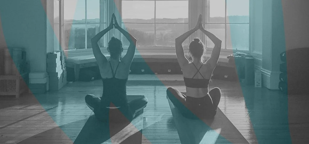 Two women practicing yoga in a sunlit room sitting on yoga mats with hands in prayer pose above their heads.