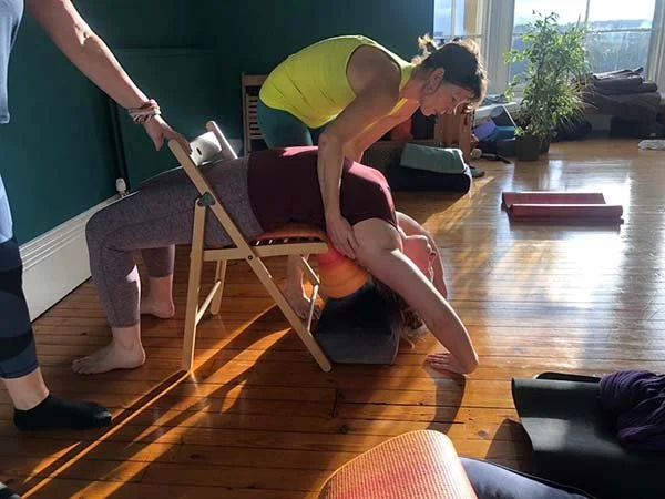 Two women participating in a chair yoga practice, with one woman leaning forward onto a chair while the other assists her with stretching over the chair in a bright yoga studio.