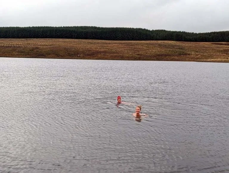 Two people swimming in a Scottish Loch with a grassy hill and forest in the background.