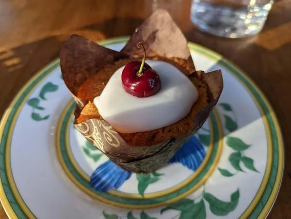A cupcake topped with white frosting and a cherry, sitting in a decorative wrapper on a patterned plate with a glass of water in the background.