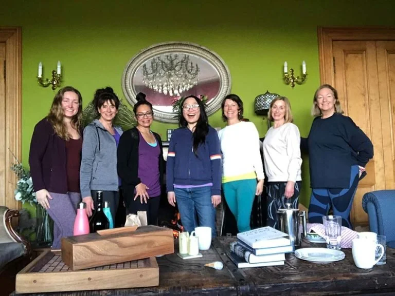 Seven women standing together in a living room, smiling. There is a large mirror, chandelier, wooden door, and green walls behind them. A table in front has books, mugs, and bottles.