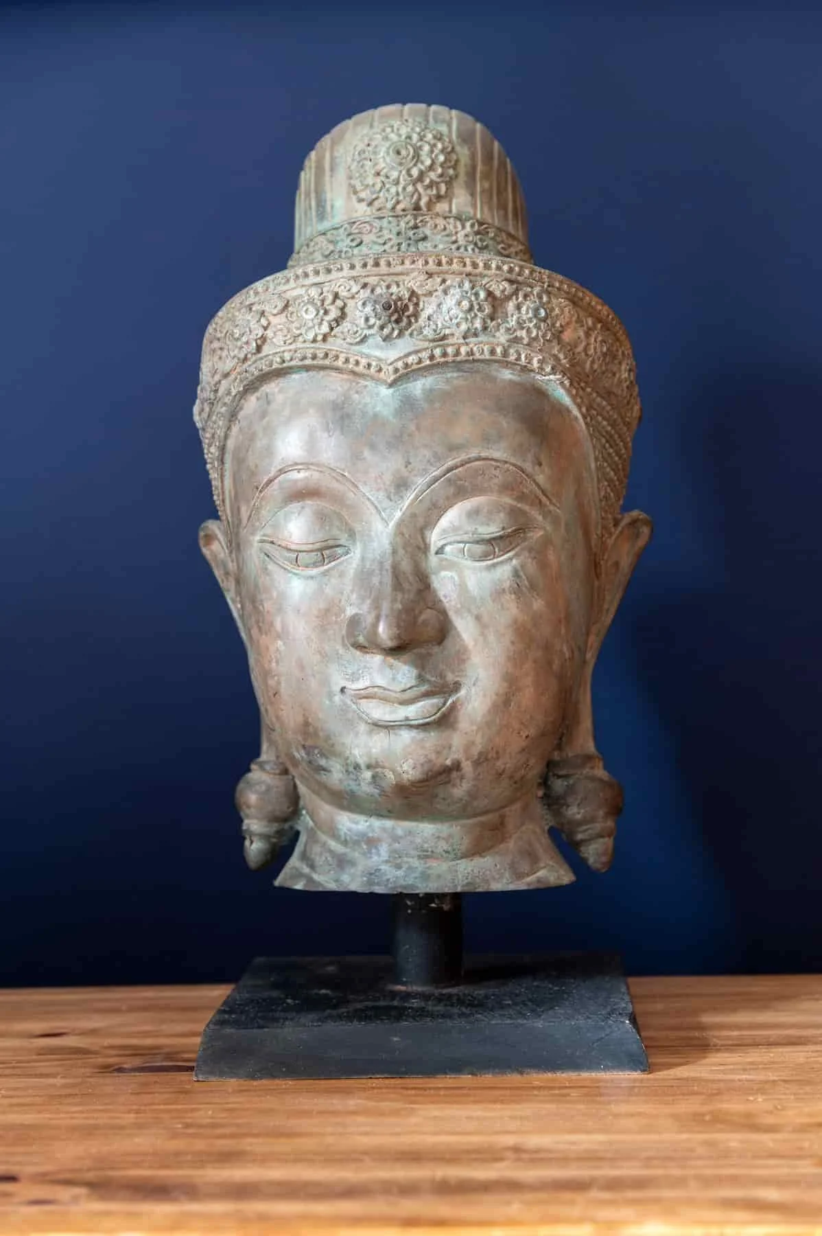 Ancient sculpture of a serene female face with intricate headdress, mounted on a stand, against a dark blue background.