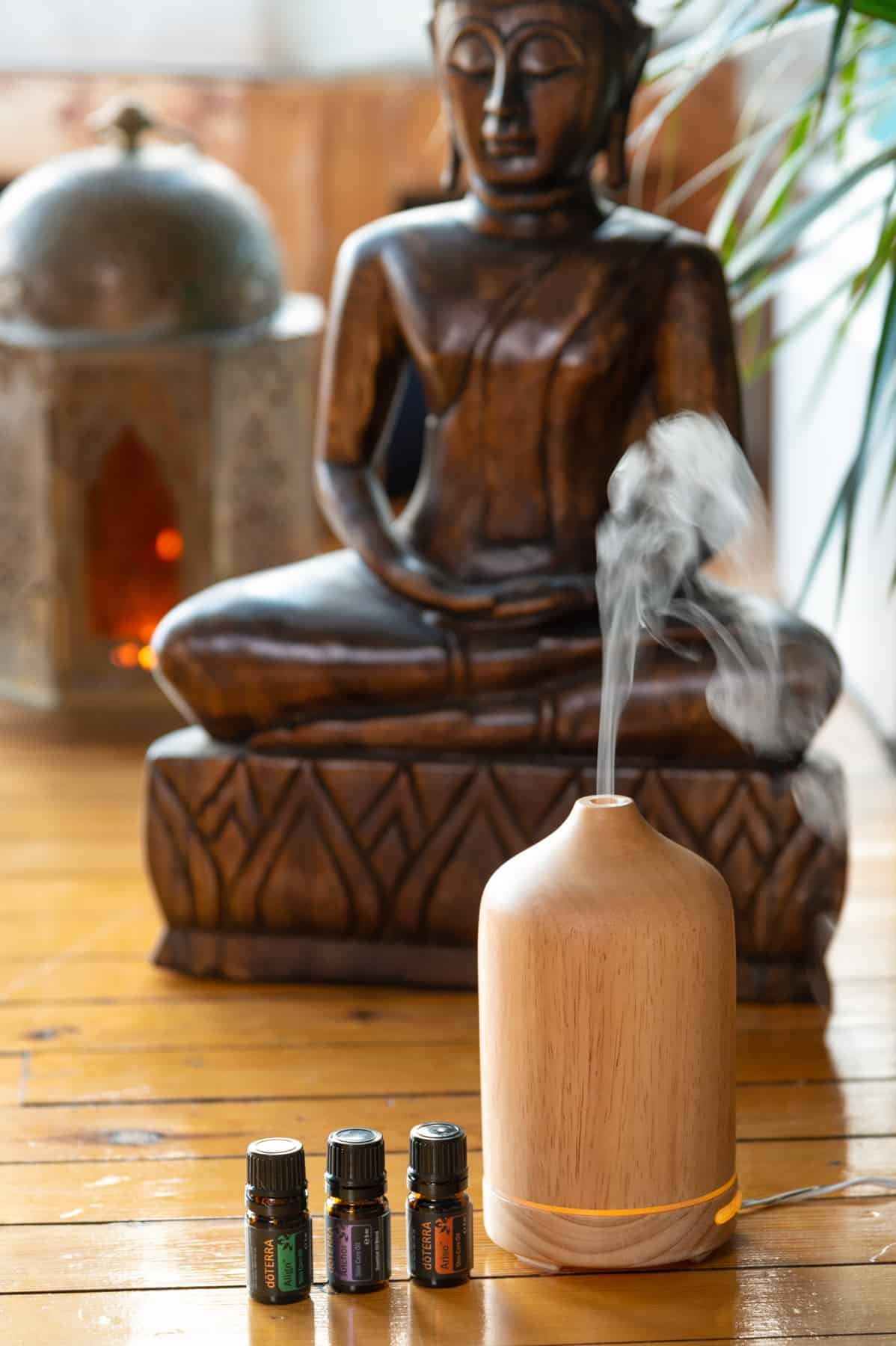 A wooden incense diffuser emitting smoke, with three essential oil bottles in front, and a wooden statue of a meditative figure in the background.
