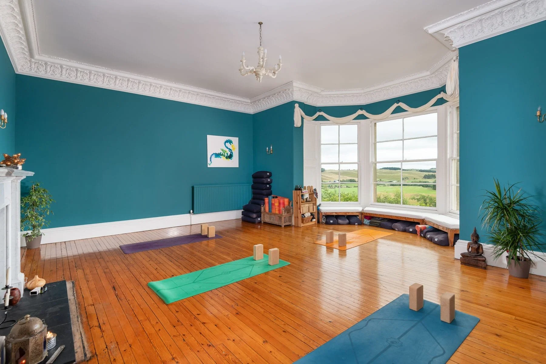 A spacious yoga or meditation room with hardwood floors and teal walls. Large bay window with white trim offers a scenic outdoor view. The room contains yoga mats, blocks, and cushions arranged for practice, and is decorated with plants, artwork, and a white chandelier.