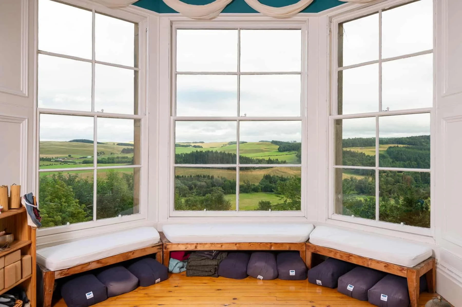 A cozy window nook with built-in benches and cushions, overlooking a scenic countryside landscape with rolling hills and scattered trees.