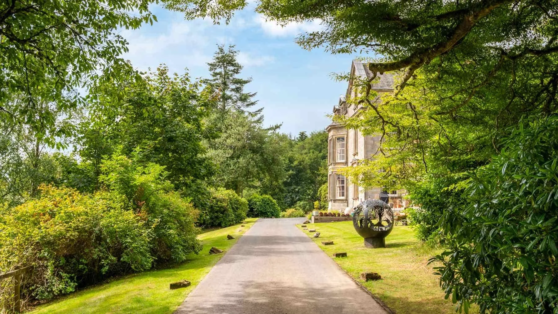 A paved pathway leading to a historic building, surrounded by lush green trees and bushes, with a decorative sculpture on the right side.