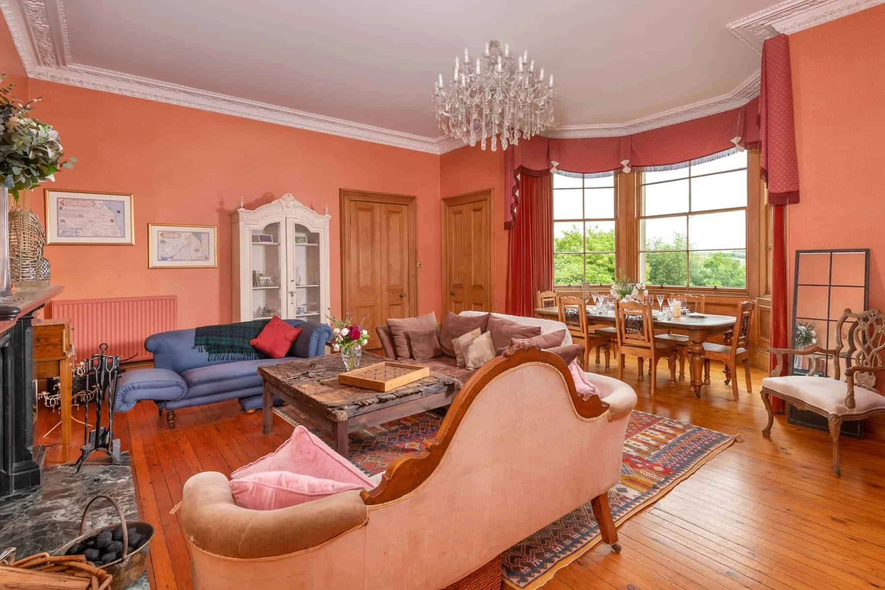 Living room with pink walls, wooden flooring, vintage furniture, large bay window, chandelier, and decorated with plants and framed pictures.