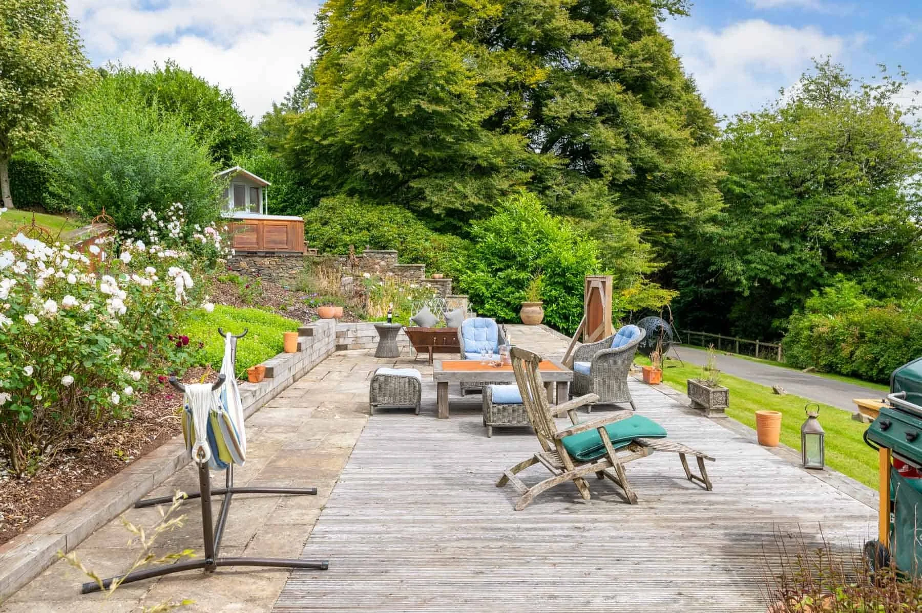 Outdoor patio with wooden deck, wicker and cushioned chairs, a table, potted plants, garden, trees, and a road in the background.
