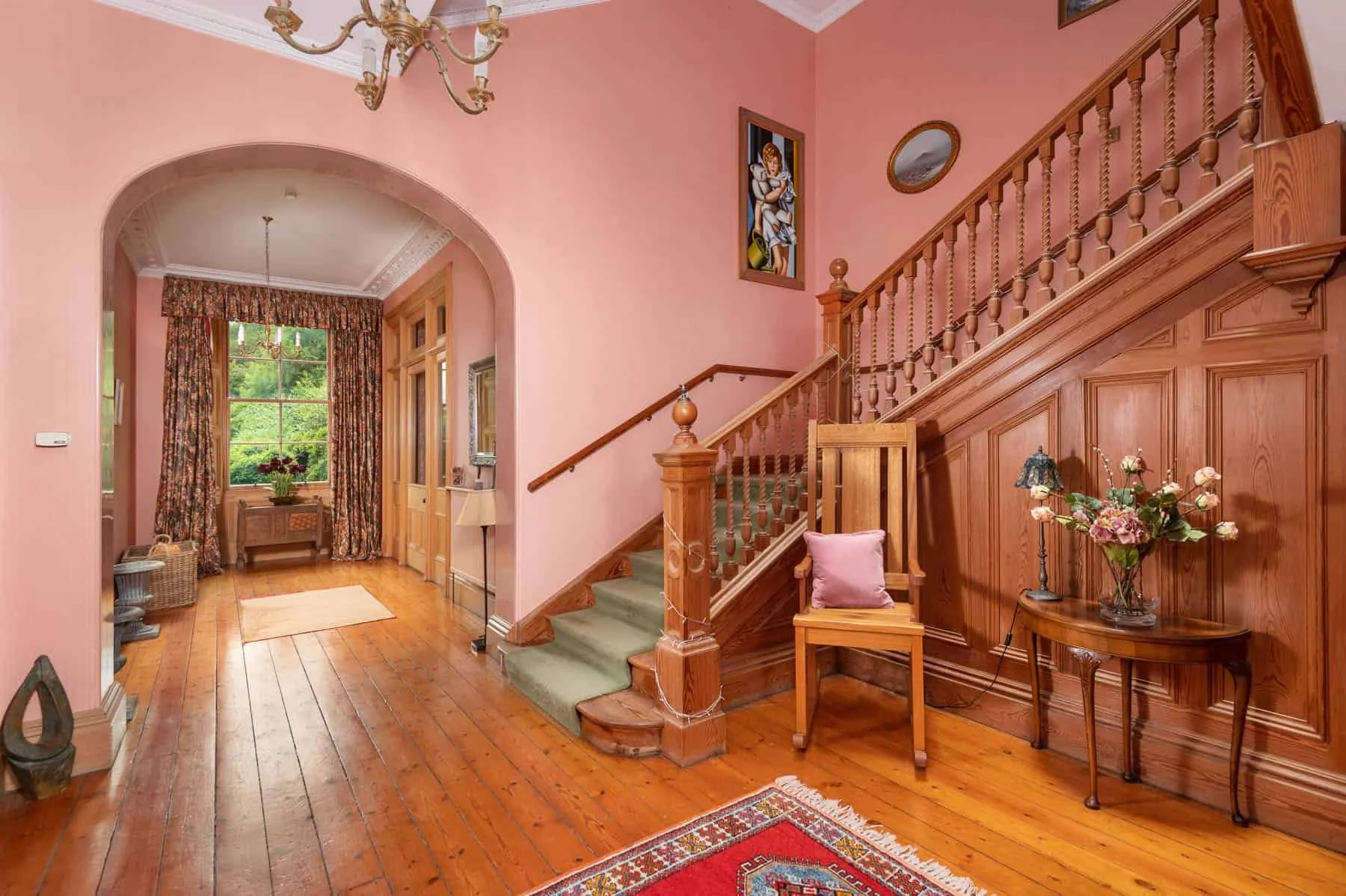 Interior of a house with a pink wall, wooden staircase, a wooden table with a flower arrangement, and a wooden chair with a pink cushion.
