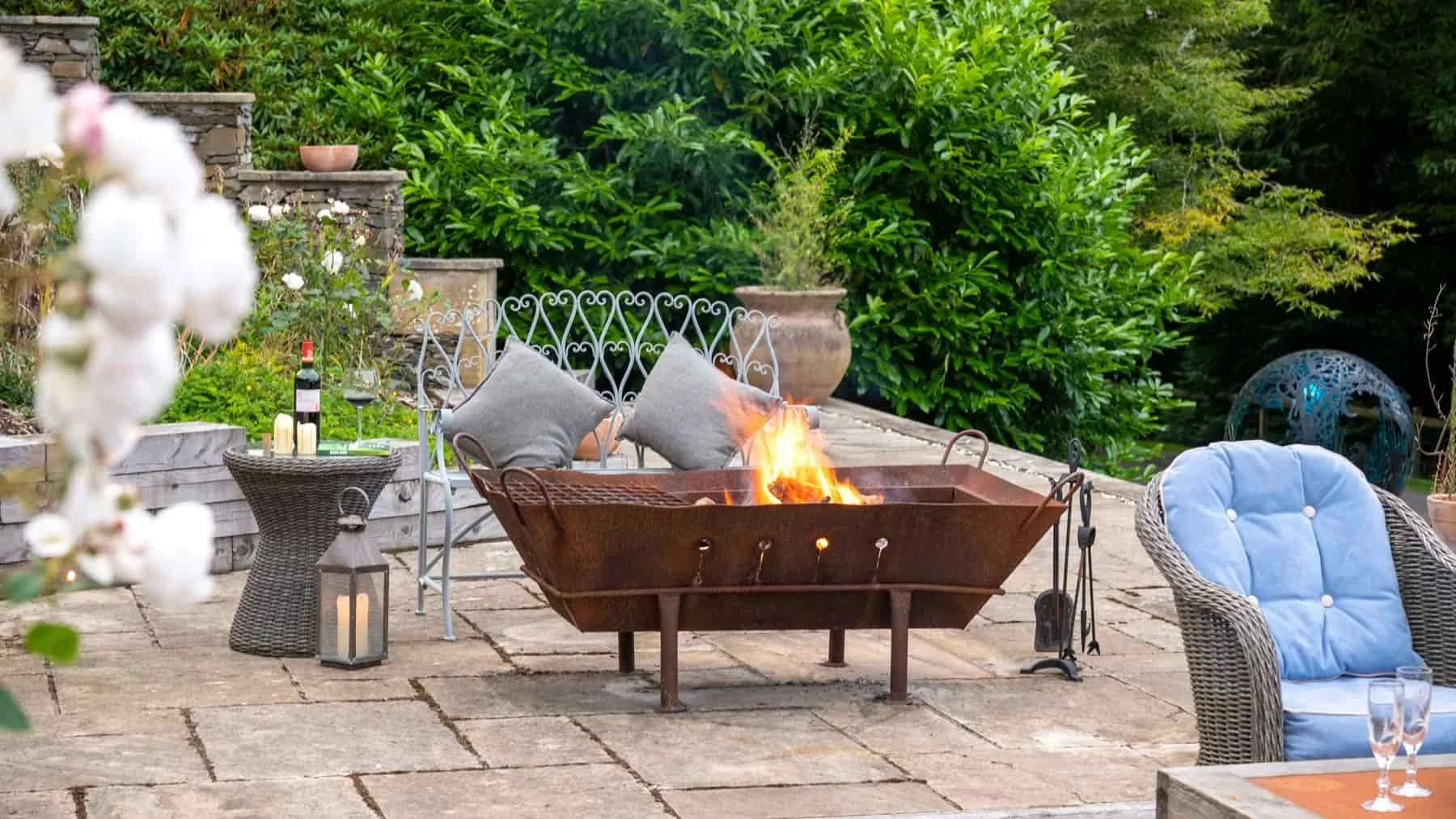 Outdoor patio with a fire pit, seating area with gray and blue cushions, lush green bushes and trees in the background, wine bottle and glasses on a small table, lanterns, candles, and various decorative items.