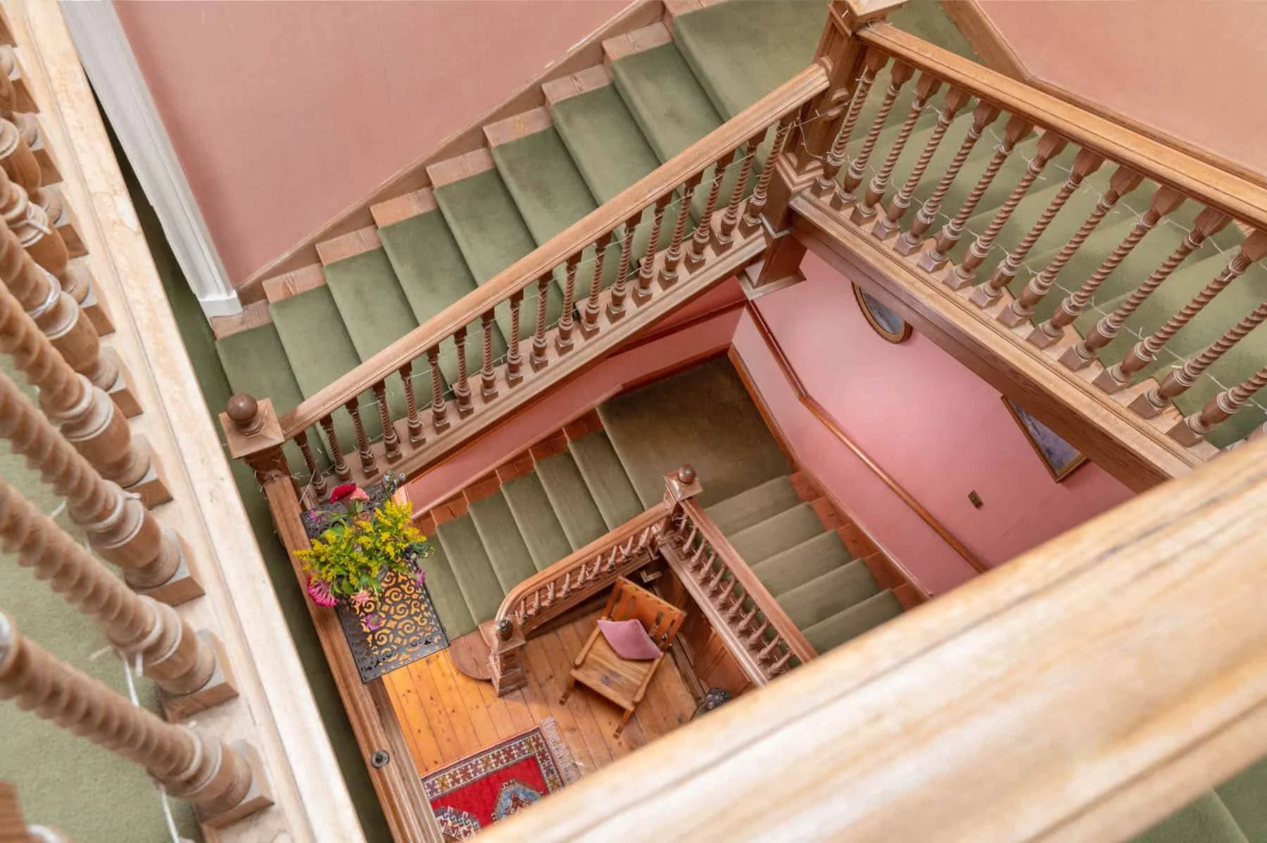 Top-down view of a wooden staircase with green carpeted steps, pink walls, and a small landing area decorated with a potted plant, a chair, and a picture frame.