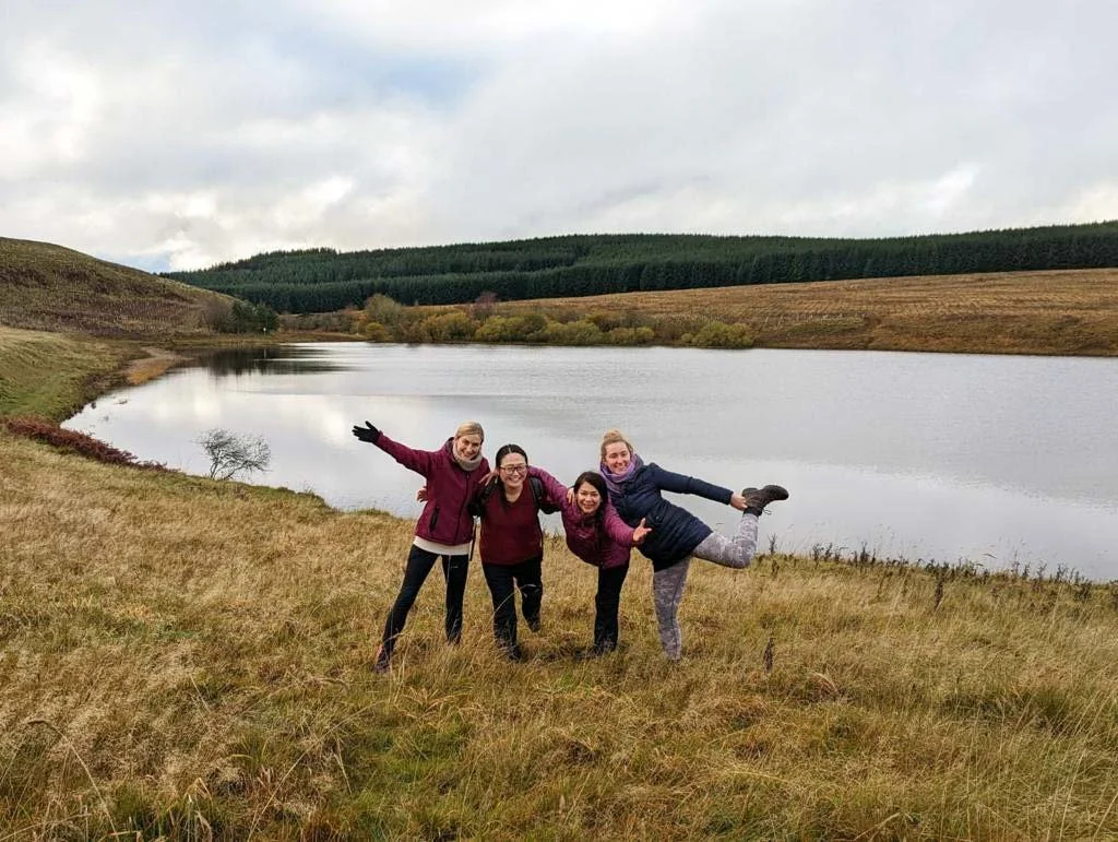 Four women standing on a grassy field near a lake, smiling and posing with arms raised and lifted legs, with hills and trees in the background on a cloudy day.