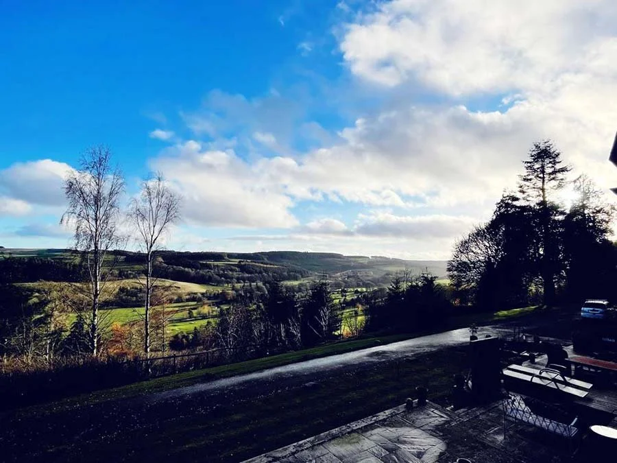 Scenic landscape view of rolling hills, trees, and a partly cloudy sky, seen from a patio with outdoor furniture.
