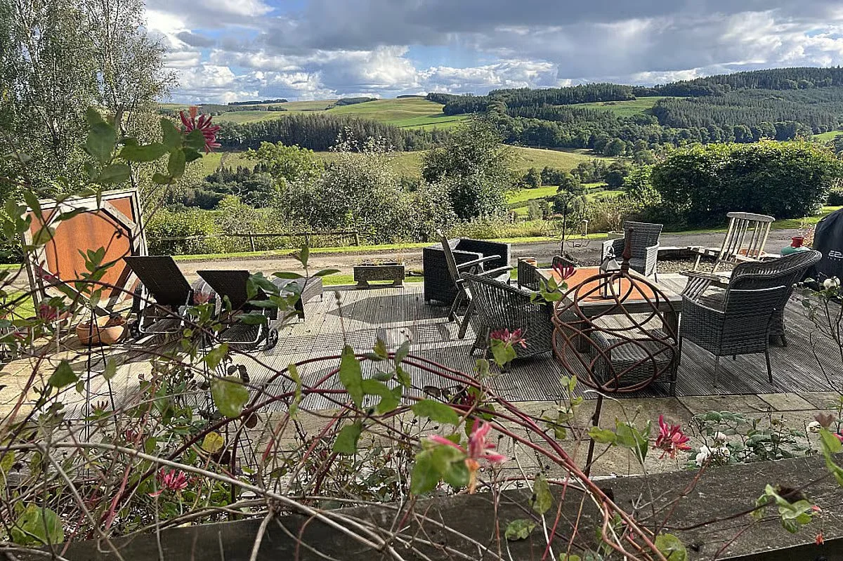 An outdoor patio with patio furniture and a scenic view of rolling green hills and trees under a partly cloudy sky.